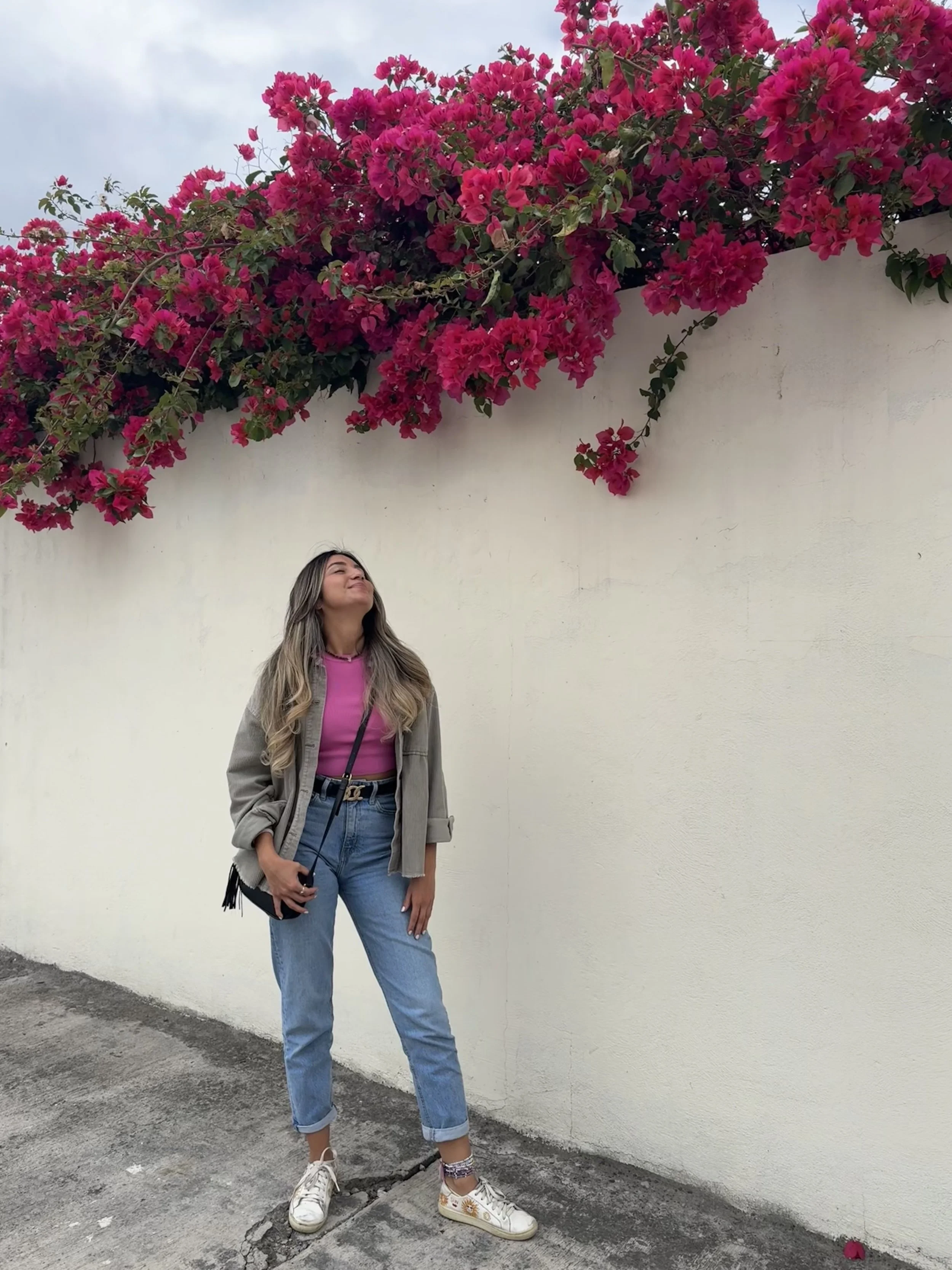 Woman standing on sidewalk in casual clothing, looking up at blooming pink bougainvillea flowers against a white wall.