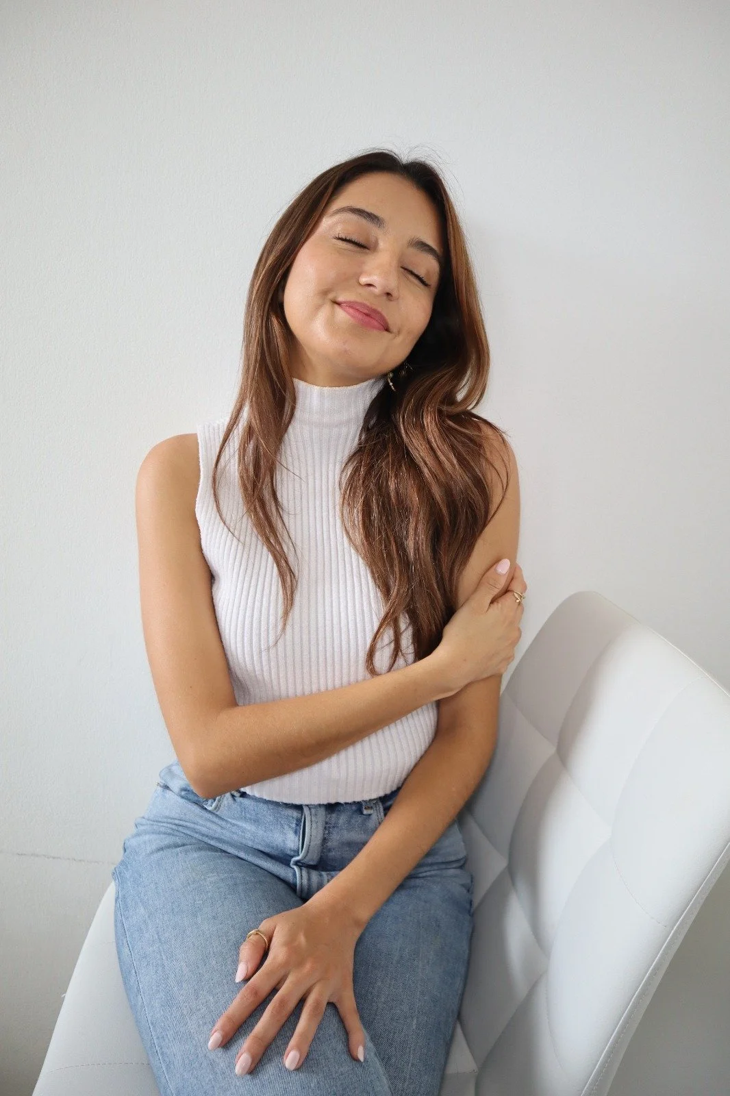 A young woman with long brown hair, wearing a white sleeveless turtleneck and light blue jeans, sitting on a white chair against a plain white wall, with her eyes closed and a gentle smile, resting her left hand on her lap and her right hand on her shoulder.