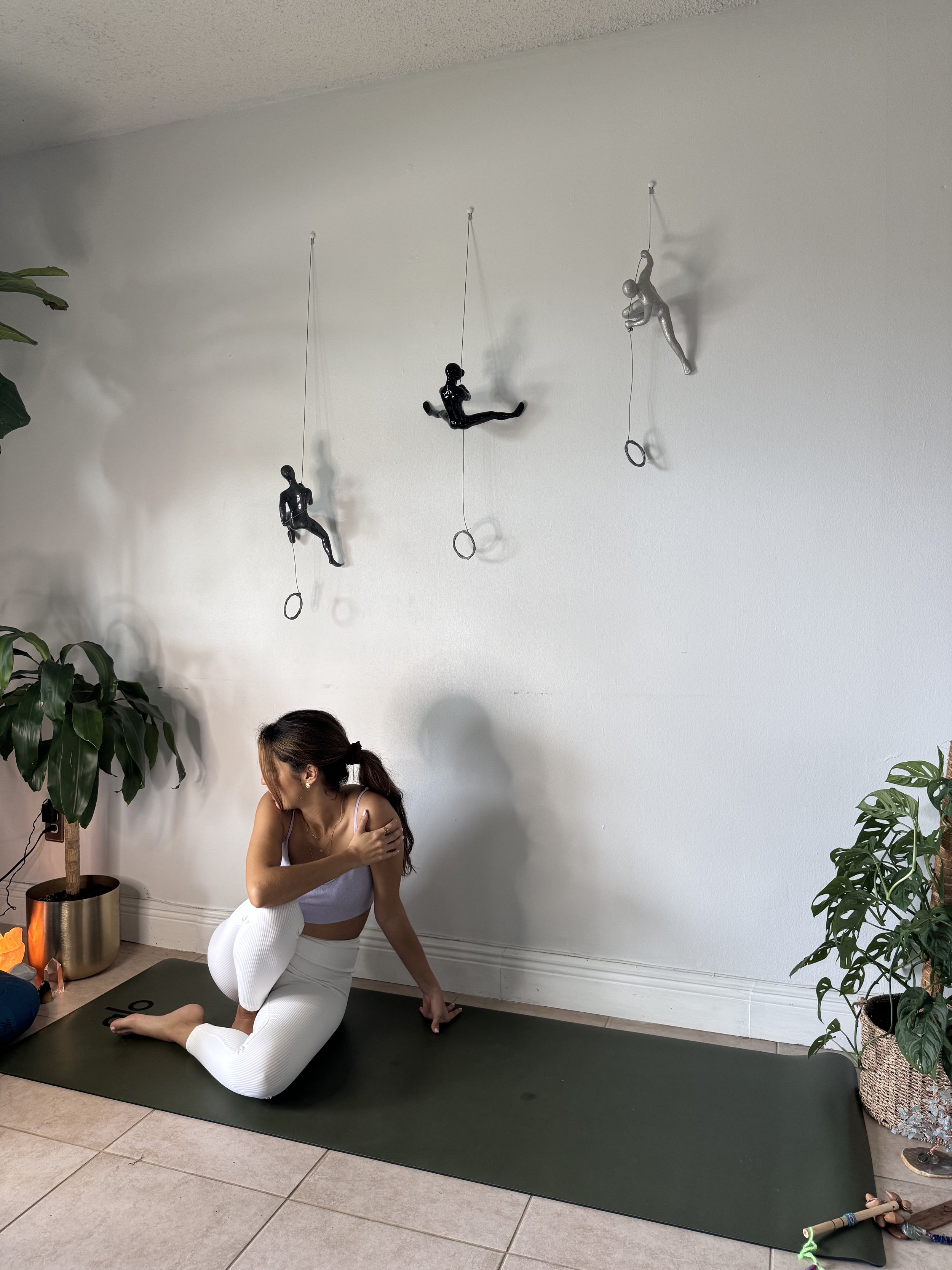 A woman practicing yoga indoors on a black yoga mat, with decorative wall hangings resembling figures hanging from the ceiling and potted plants on either side.
