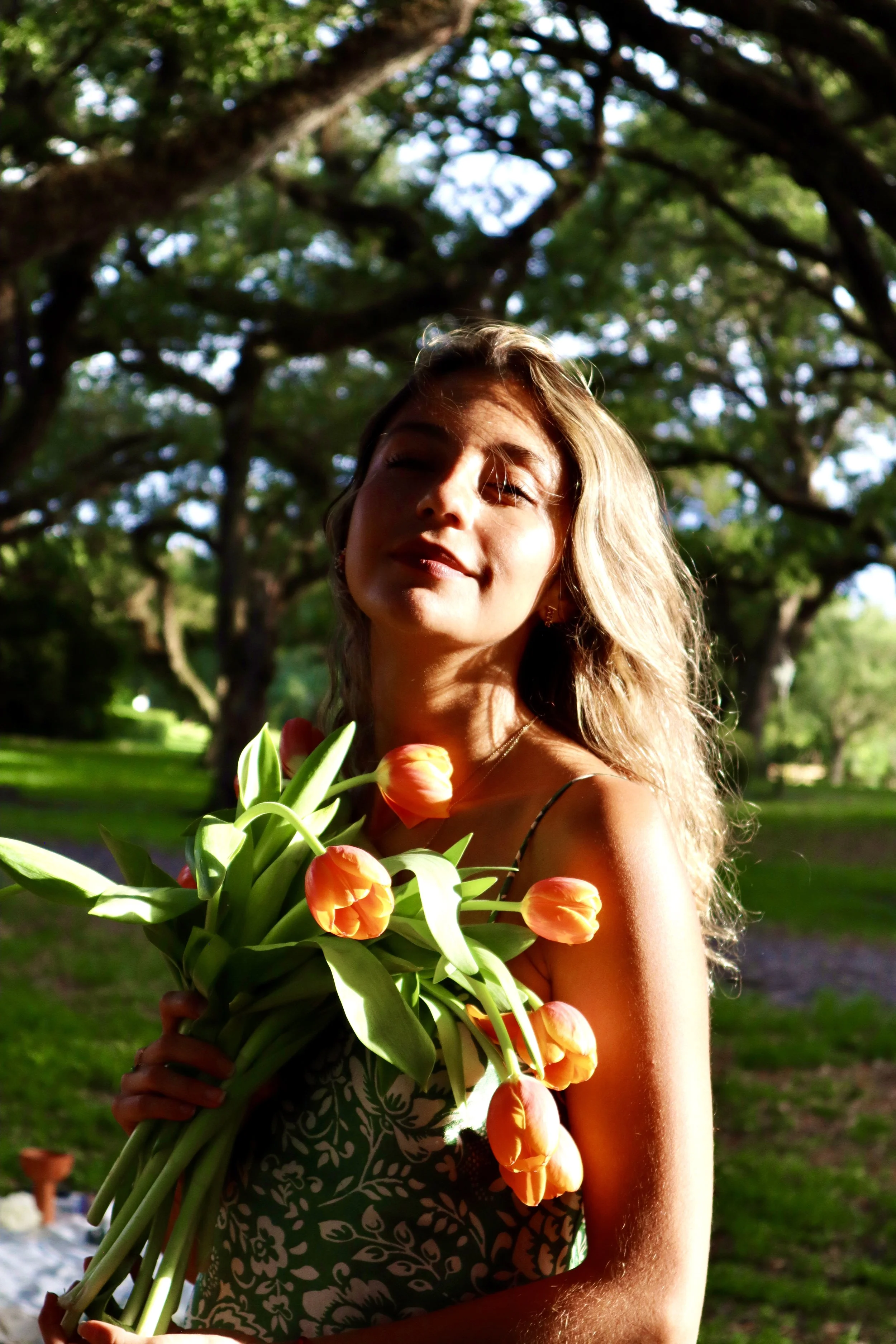 Paola amezquita UKUMANTA Miami  with light skin and blonde hair holding a bouquet of orange tulips outdoors in a park with large trees and green grass.