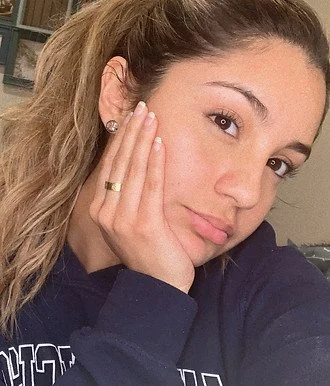 Close-up of a young woman with light brown hair, resting her face on her hand, wearing a gold ring and earrings, dressed in a navy blue hoodie.