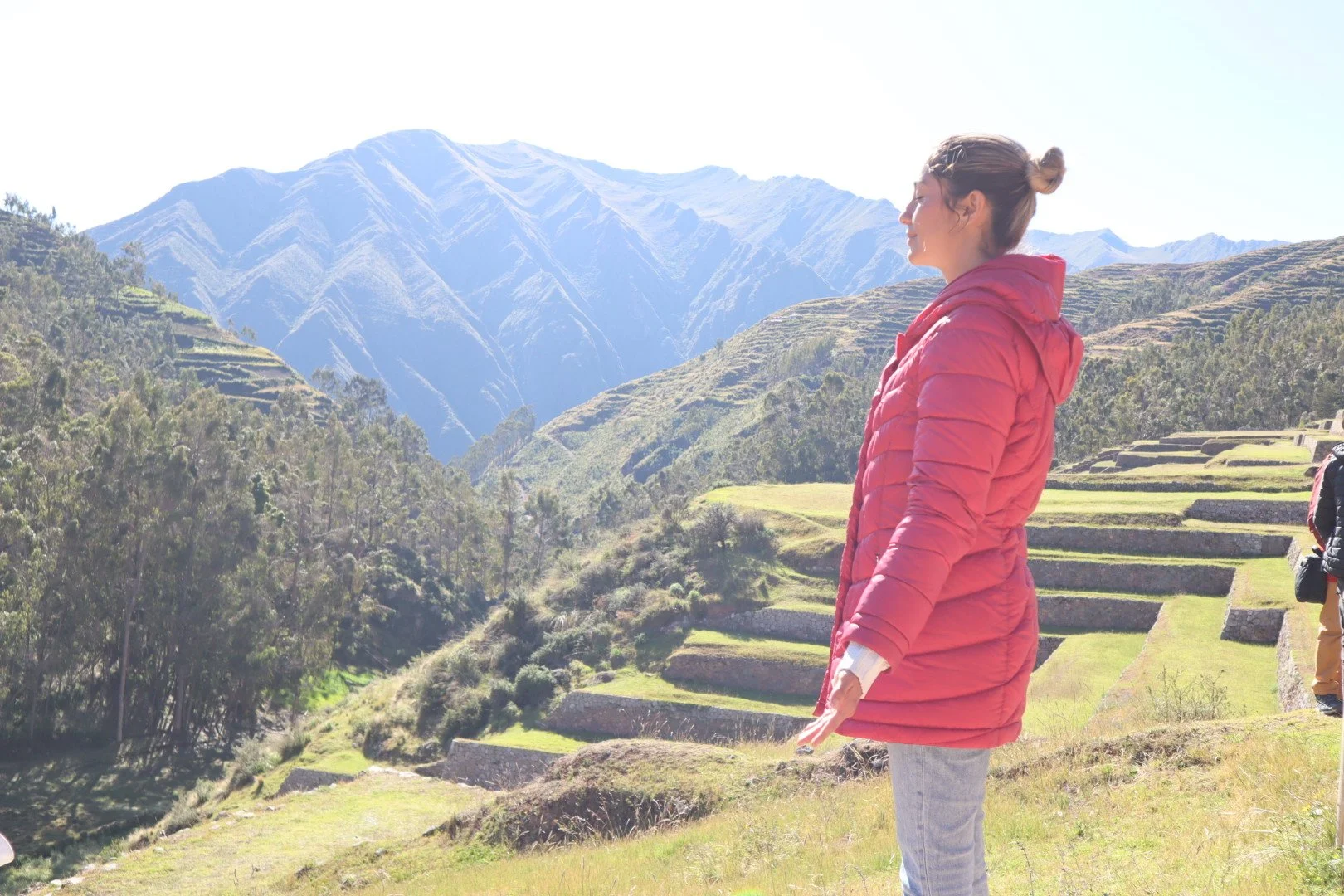 Paola Amezquita in a red jacket standing on a grassy hill overlooking terraced agricultural fields and mountains