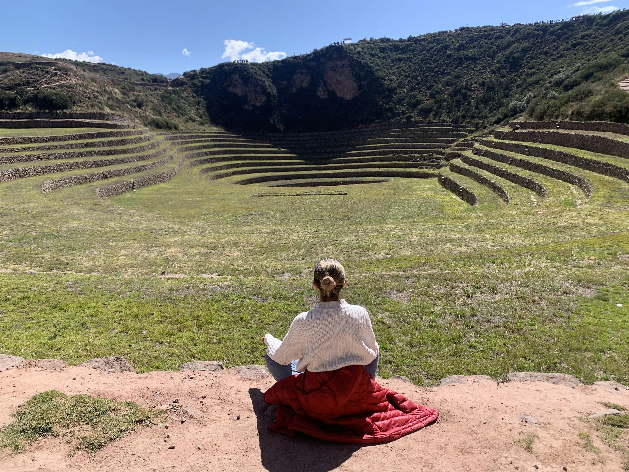 Person sitting on the ground facing an ancient stepped amphitheater or archaeological site with terraced rock walls, located in a mountainous area under a clear blue sky.