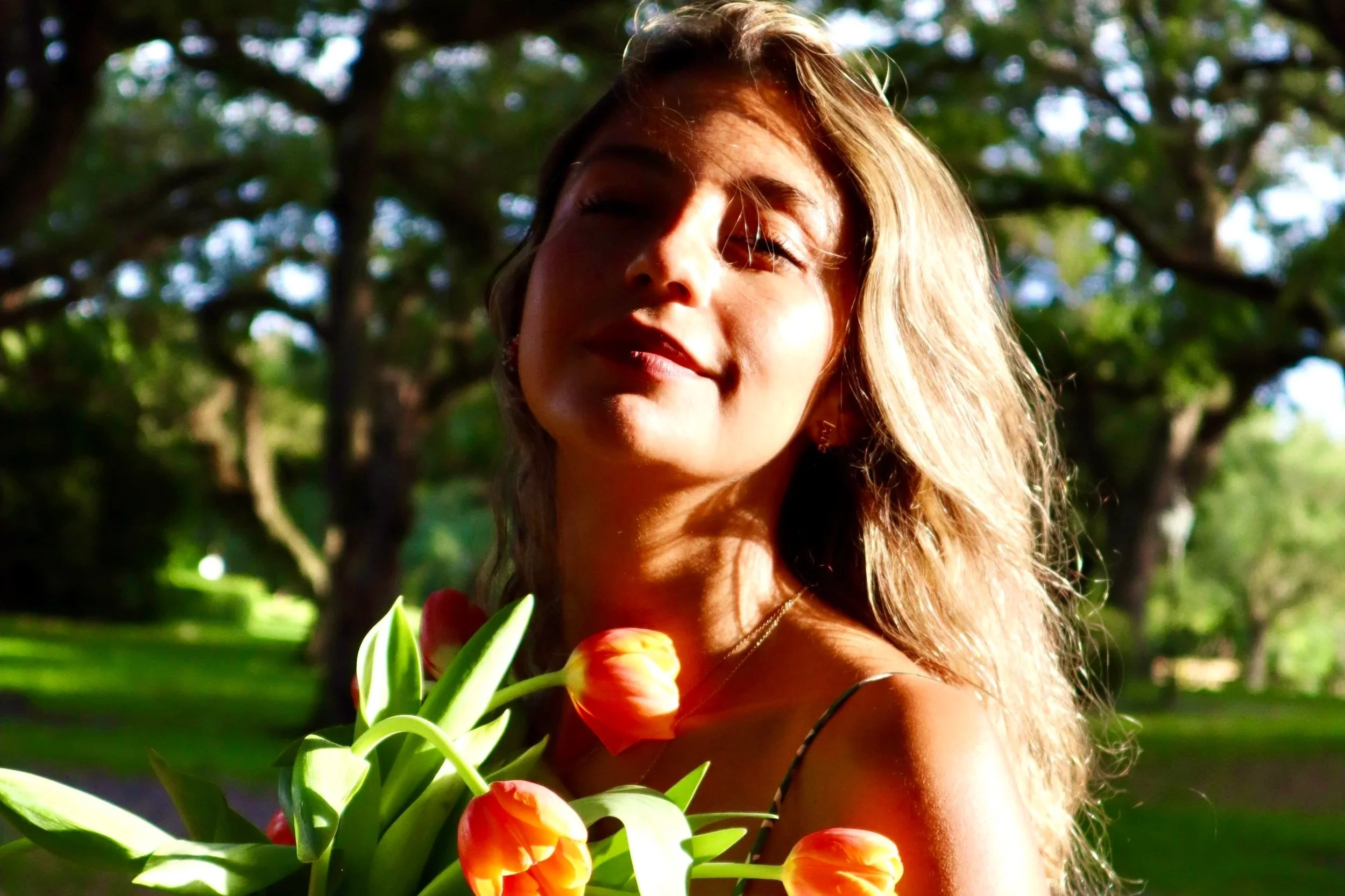 A woman with long curly hair holding a bouquet of orange tulips outdoors in a sunny park with trees in the background.