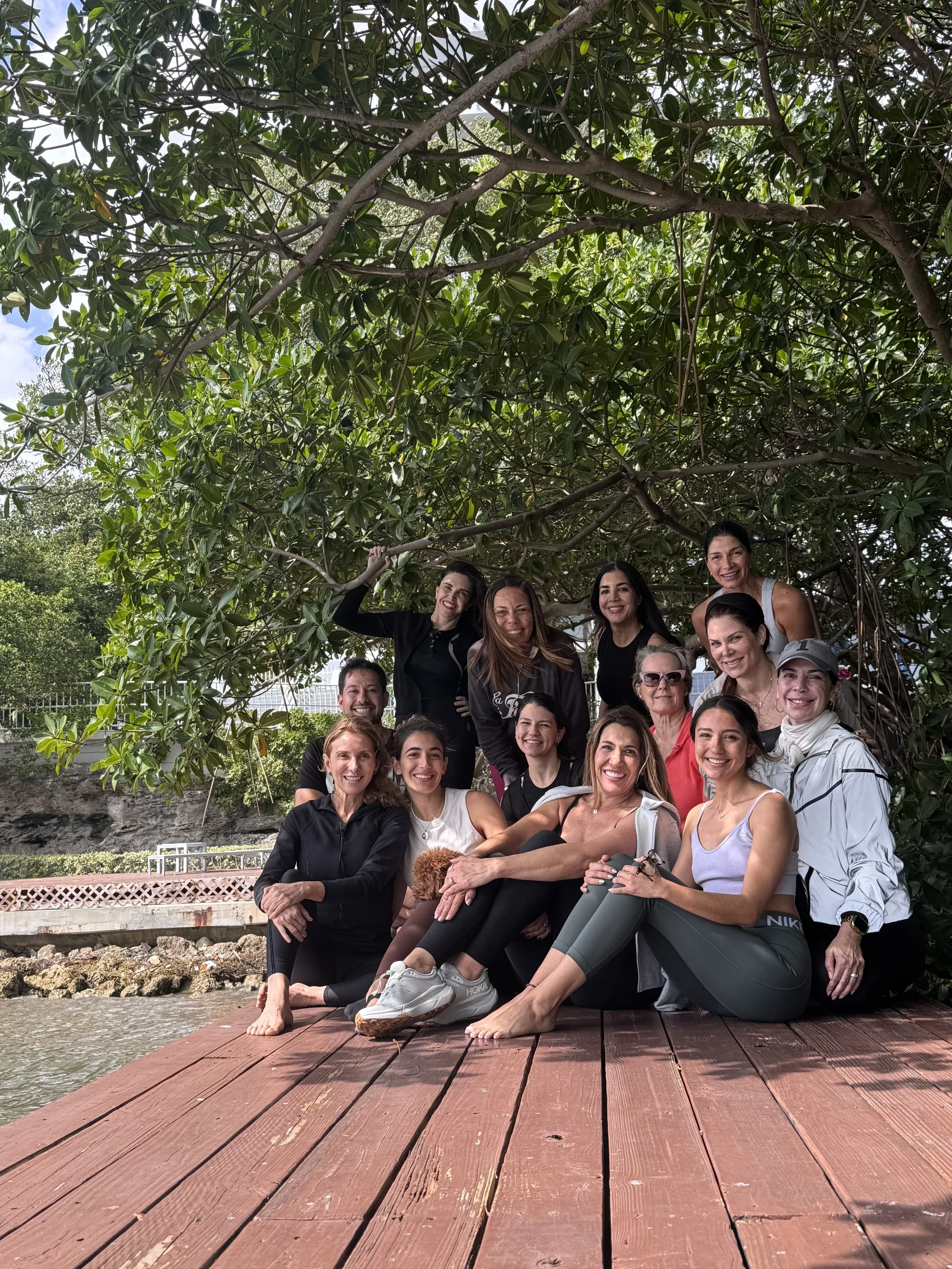 Group of women sitting and standing on a wooden dock under a large tree in a natural outdoor setting, smiling and enjoying the moment.