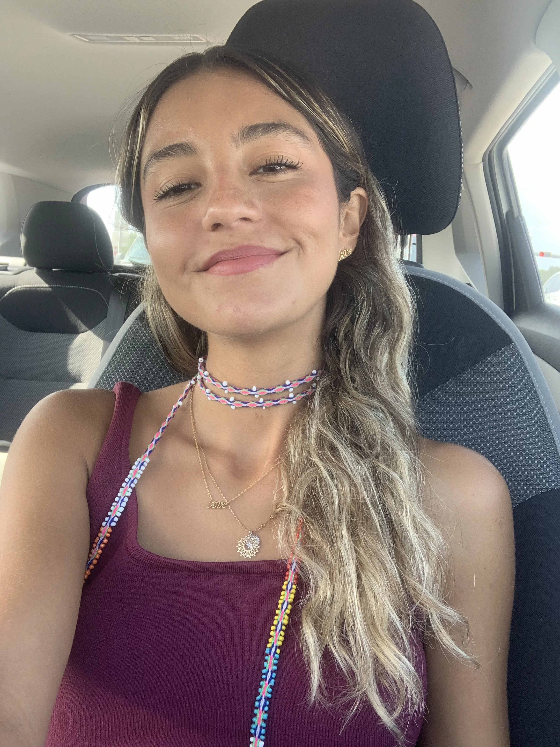 A young woman with light brown, wavy hair and a smile, sitting in a car. She is wearing a maroon tank top, colorful beaded necklaces, and a gold necklace with the word 'love.' She has stud earrings and is enjoying the moment.