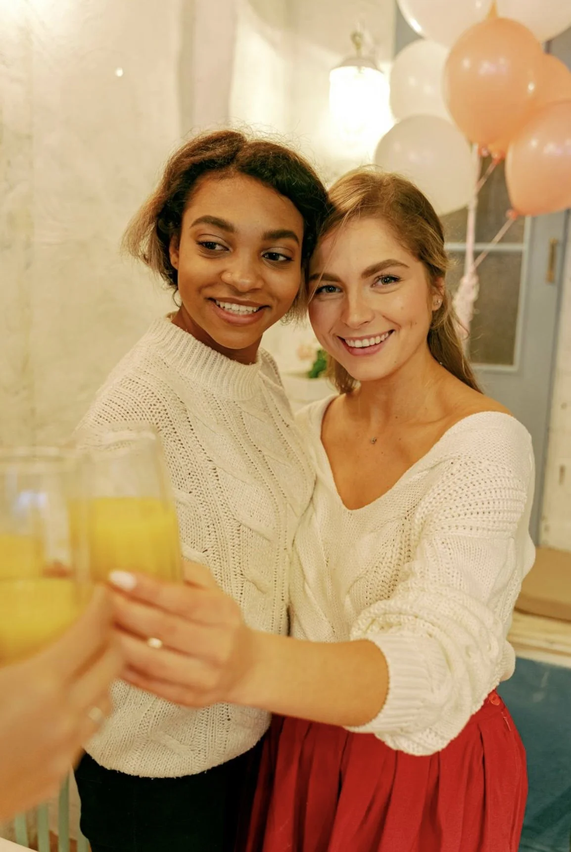 Two women smiling and holding drinks, celebrating at a gathering with balloons in the background.