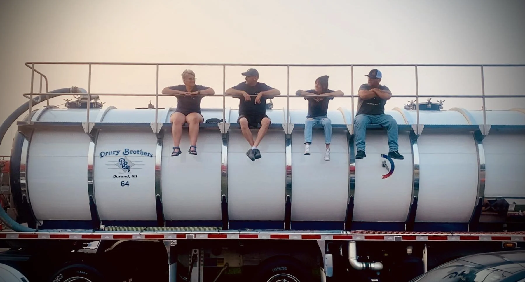 Four people sitting on top of a large white tank truck, each with their arms crossed, in Durand, MI, with a clear sky behind them.