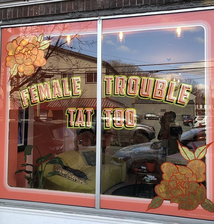 Storefront window with signs that read "Female Trouble Tattoo" in colorful letters, decorated with floral designs on the corners, reflecting a street with houses and cars.