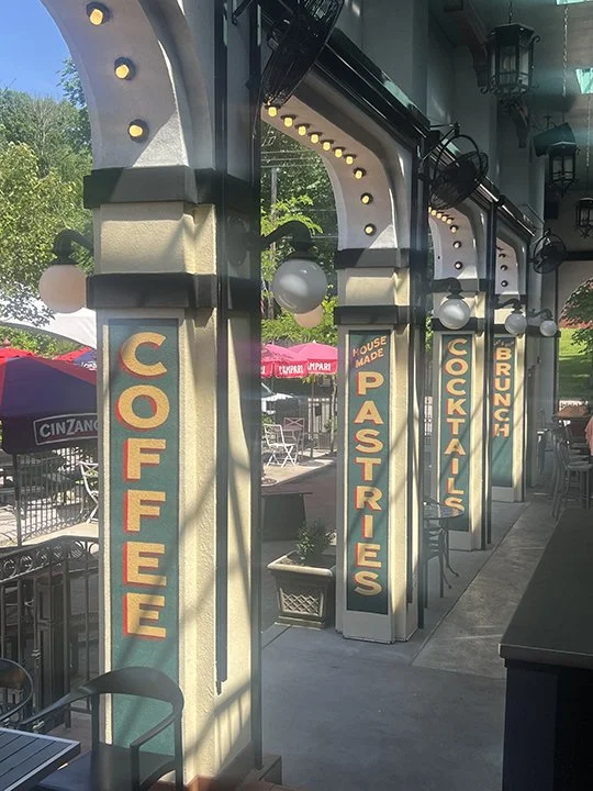 Outdoor restaurant patio with columns displaying signs for coffee, pastries, house made, cocktails, and brunch under a covered roof with hanging lights.