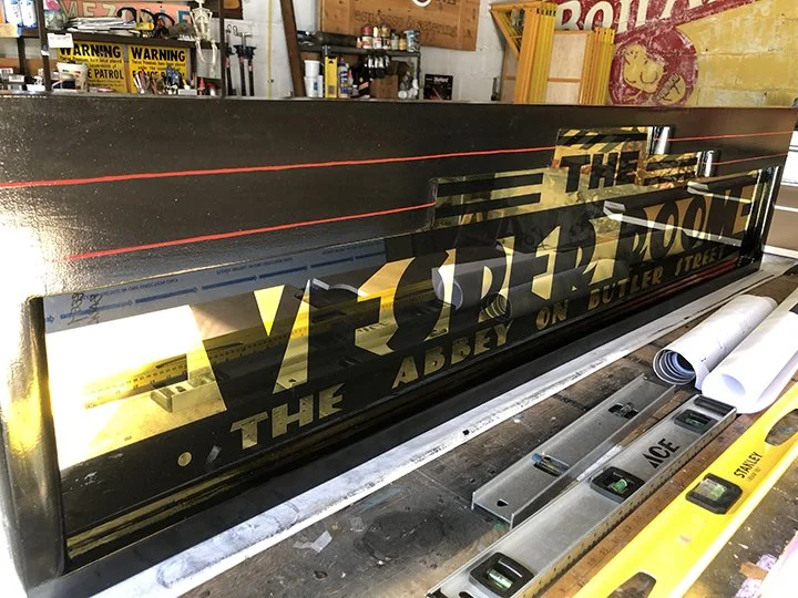 A sign or display board in a workshop, with partially visible text reading "THE VESPER ROOM" in full mirror gold leaf