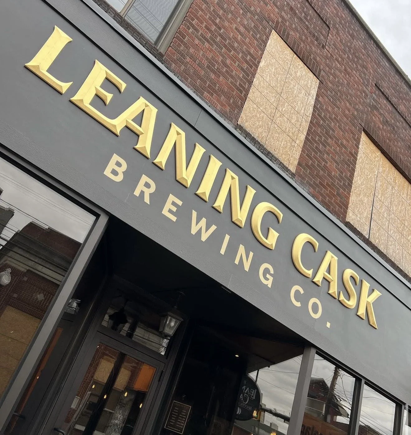 Exterior of Leaning Cask Brewing Co. building with a black sign featuring large gold lettering and windows reflecting the street.