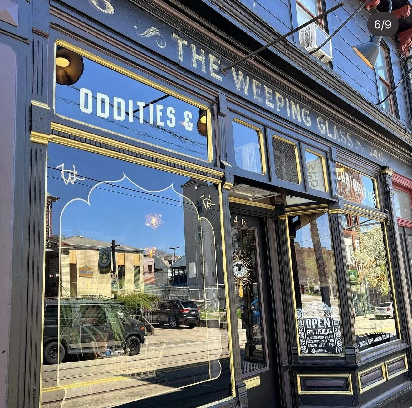 The Weepling Glass storefront with black and gold trim, displaying signs for oddities and curiosities, and an eye graphic on the door. Reflections of neighboring buildings and a street scene can be seen in the windows. The store's hours and an open s