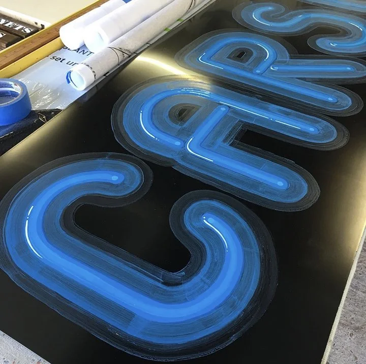 Close-up of a faux neon sign spelling 'CARSON' in blue lights on a black background, with some tools and papers nearby.