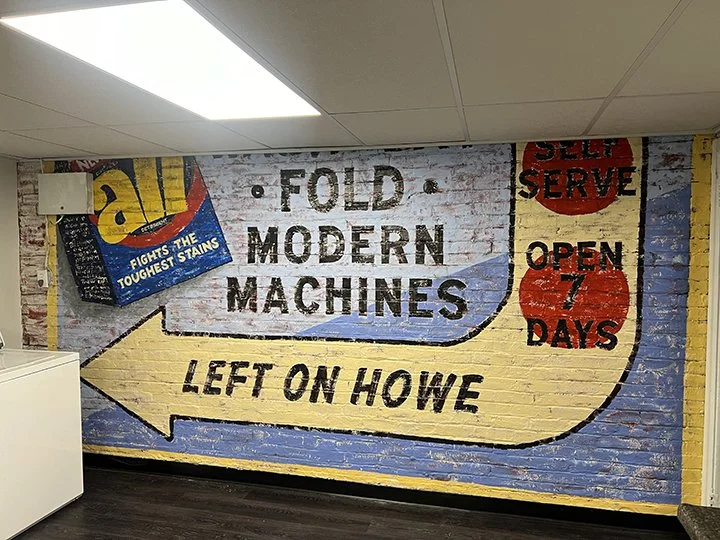 Brick wall with hand-painted advertisement for a laundry that folds, modern machines, and is open 7 days. Large yellow arrow pointing left with the words "Left on Howe."