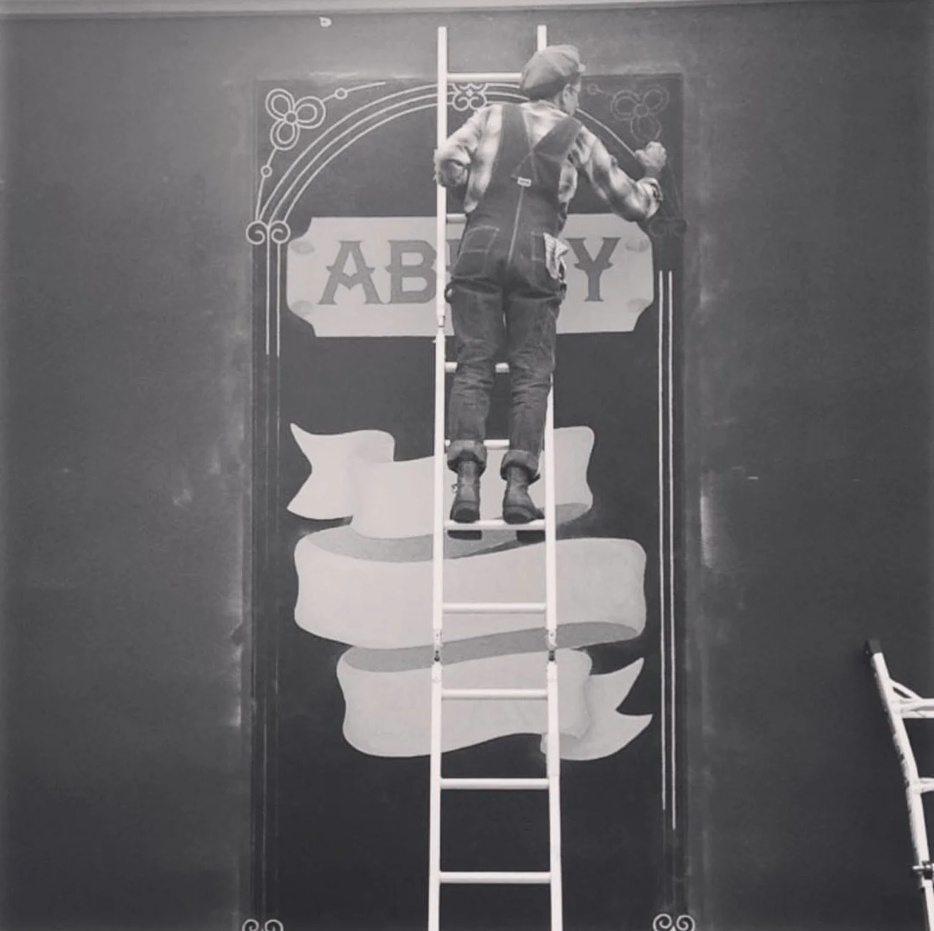 A person on a ladder painting a large sign on a wall that says 'ABBEY' with decorative designs around it.