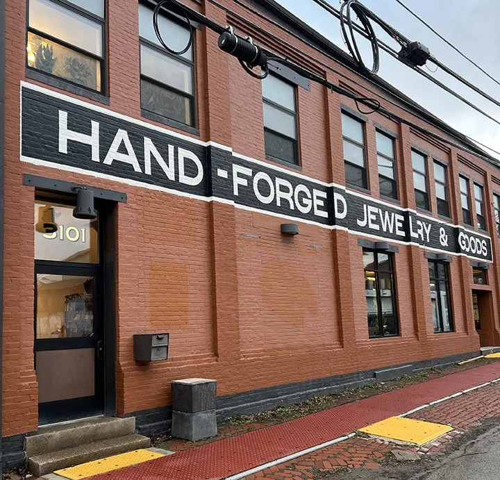Exterior of a red brick building with large black and white sign reading 'Hand-Forged Jewelry & Goods'.