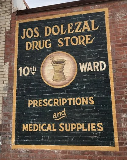A painted sign on a brick wall advertising Jos Dolezal Drug Store, located in the 10th Ward, offering prescriptions and medical supplies, with a mortar and pestle illustration in the center.
