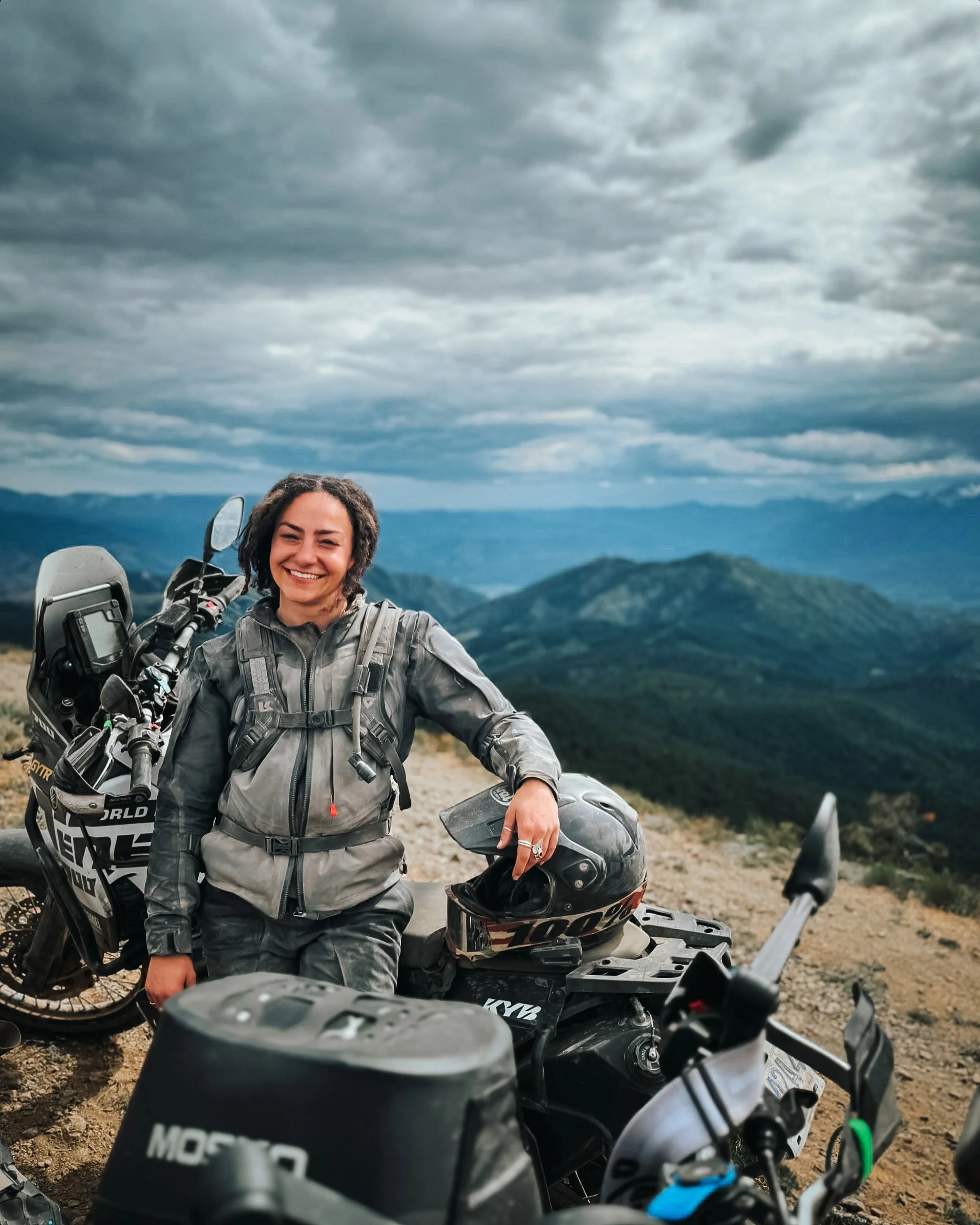 Golnoosh Namazi smiling beside her motorcycle at a mountain viewpoint under dramatic clouds.