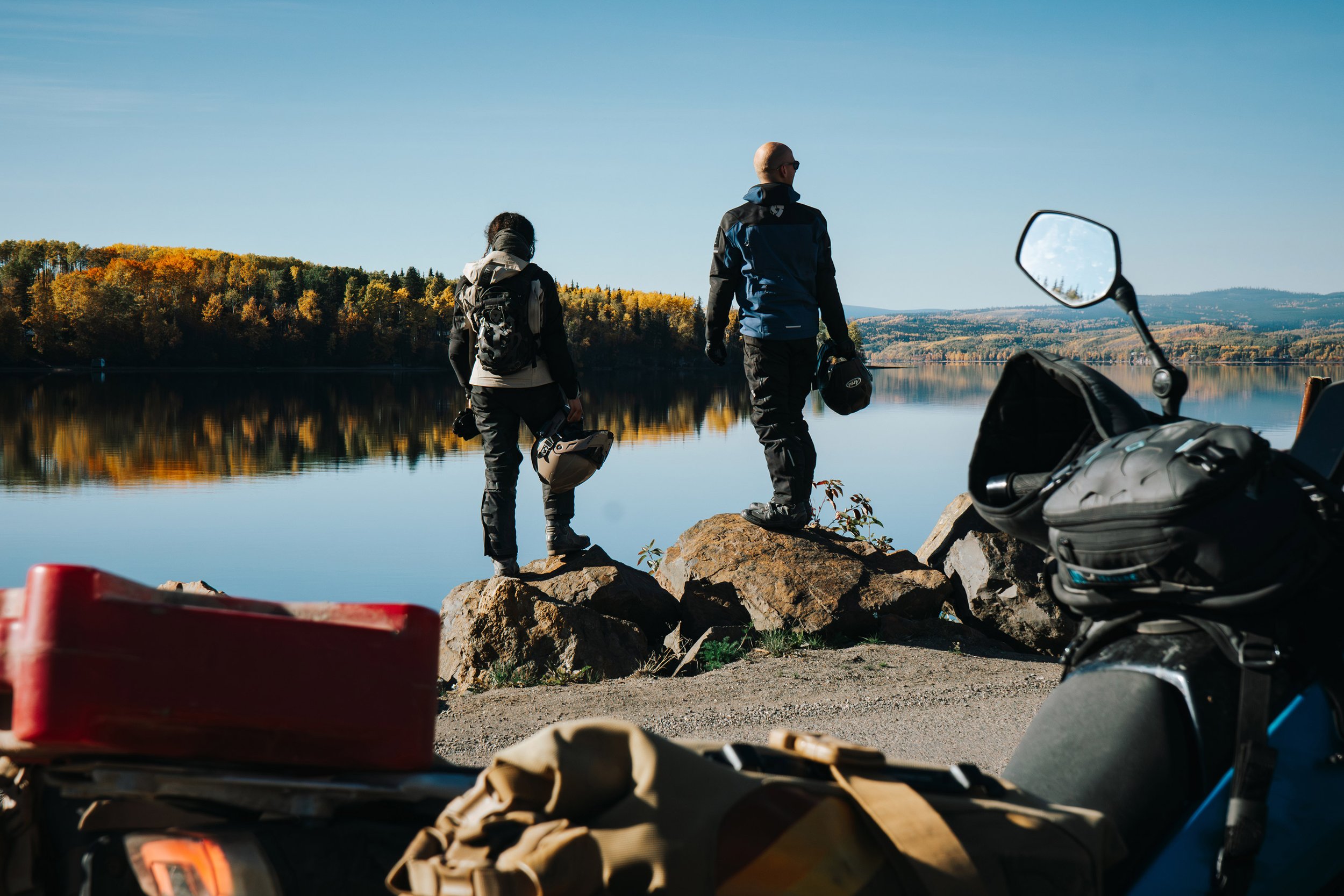 Golnoosh Namazi and a fellow rider stand on rocks at the François Lake ferry crossing near Grassy Plains, overlooking calm water during the Route 16 Backroads Adventure Guide project in Northern British Columbia.