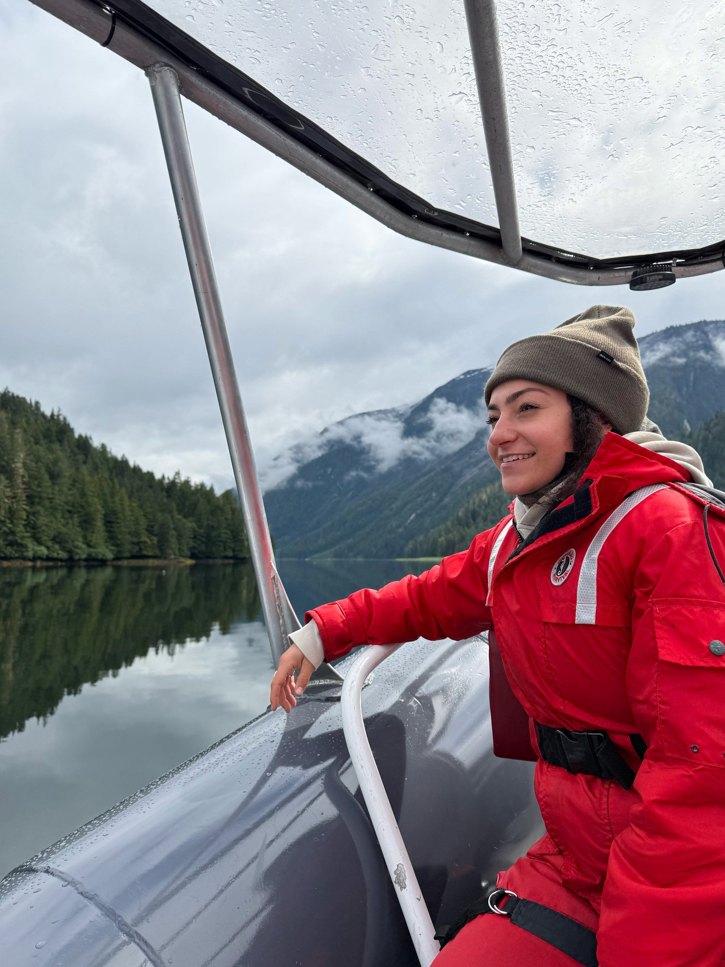Golnoosh Namazi riding in a boat on a misty fjord near Khutzeymateen Lodge, wearing red rain gear with forested mountains in the background.
