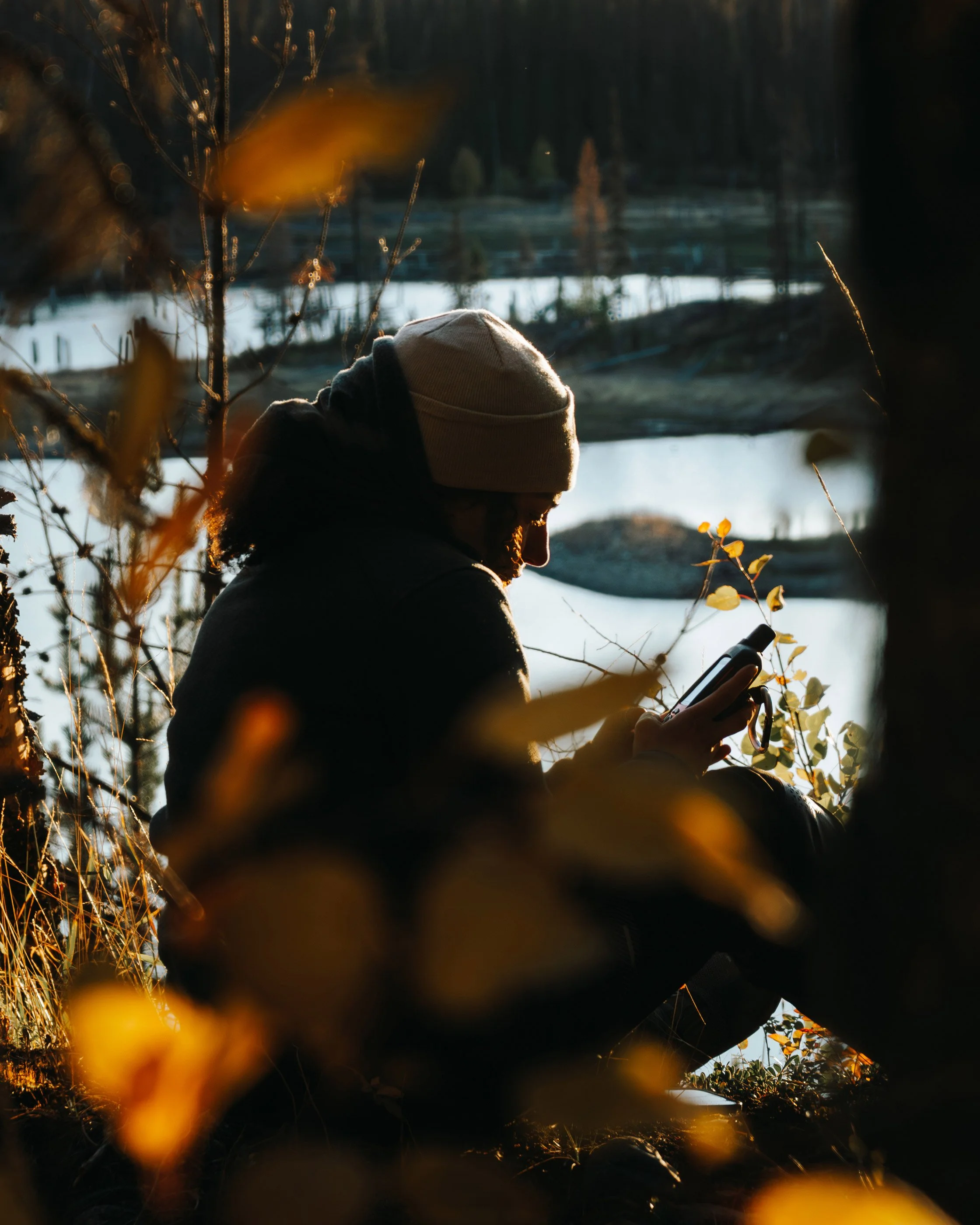 Golnoosh Namazi sitting near the water at Little Andrews Bay Provincial Park on Ootsa Lake, checking her GPS in autumn light during the Route 16 Backroads Adventure Guide Project.