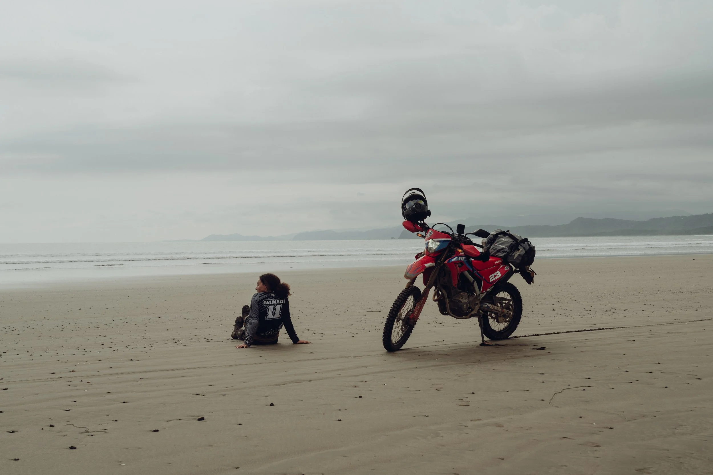 Golnoosh Namazi sitting on a wide beach beside a red dirt bike in Costa Rica during a project with Infinite Adventures.