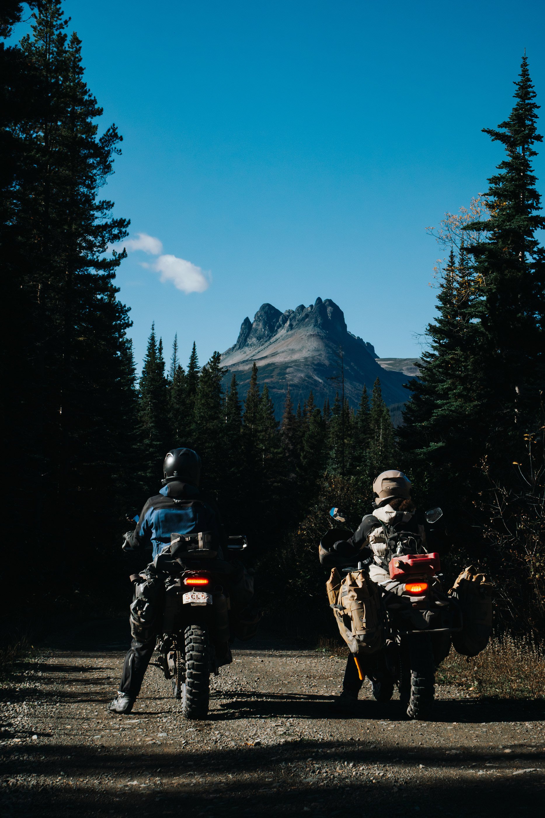Two adventure riders on loaded motorcycles stopped on a forest road, facing a jagged mountain peak in Northern BC.