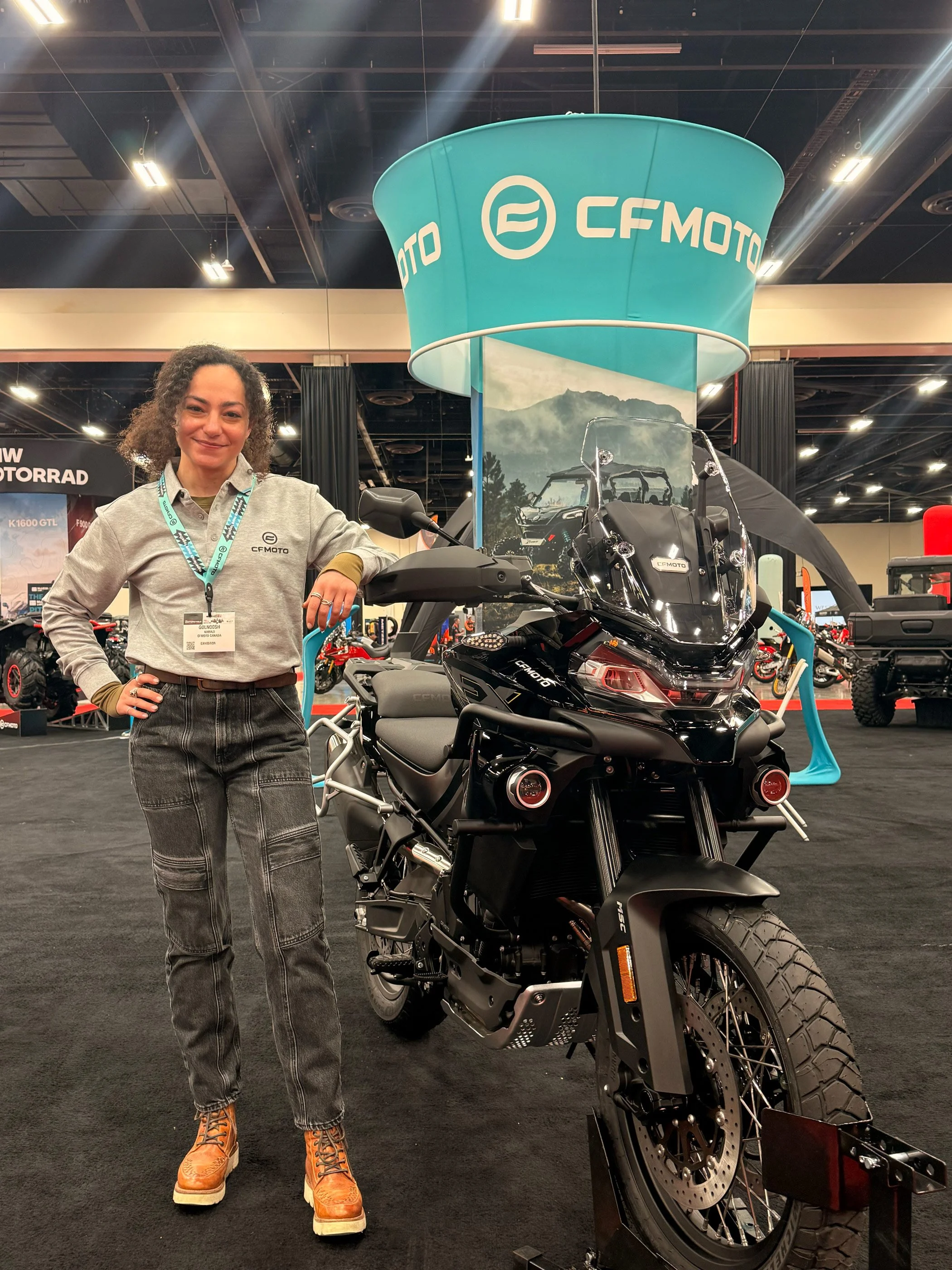 Golnoosh Namazi representing CFMOTO at a motorcycle trade show, standing beside a CFMOTO adventure touring bike at the CFMOTO booth.