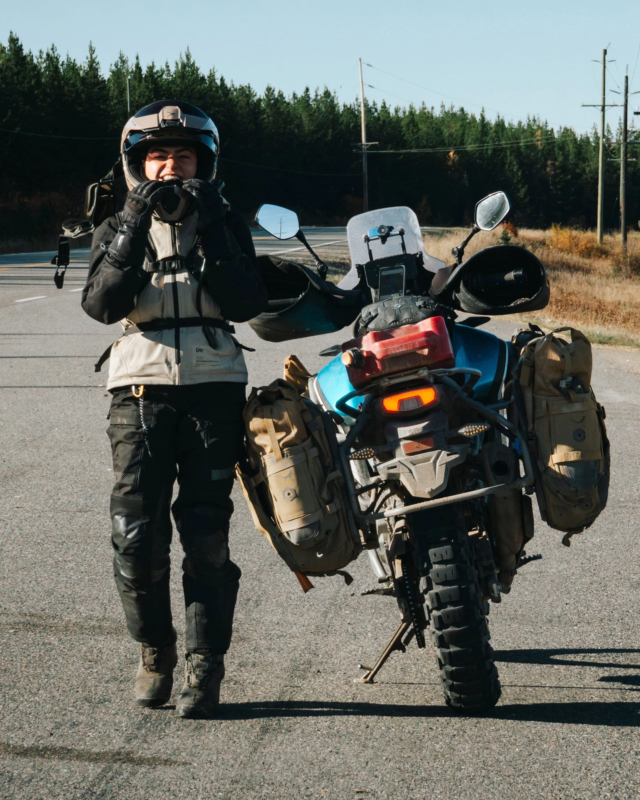 Golnoosh Namazi. Northern BC place-based storyteller & adventure rider standing beside a loaded motorcycle on a rural road.
