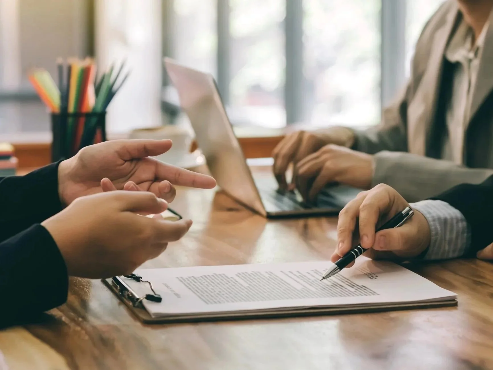 Two people discussing or reviewing a document on a clipboard while another person uses a laptop in the background at a wooden table with office supplies.