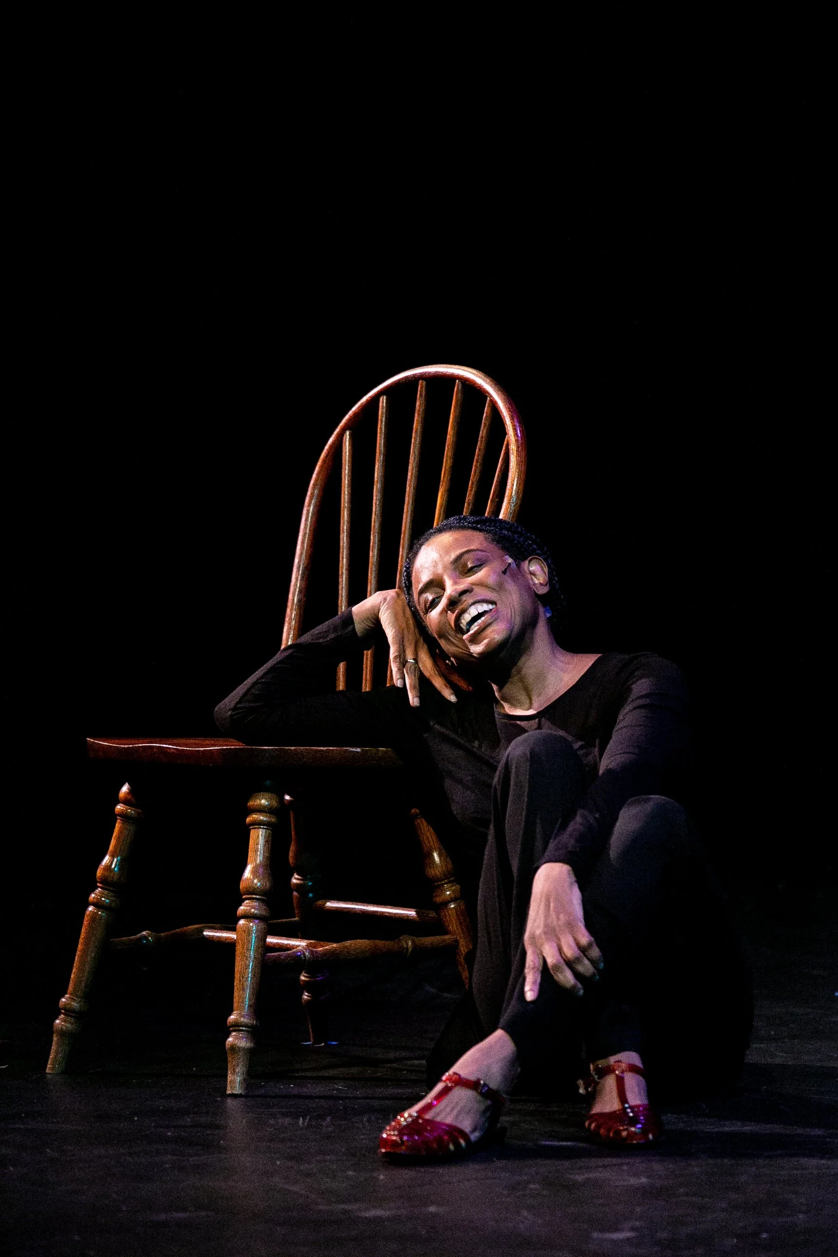 A woman singing or acting on stage next to a wooden chair, dressed in black with red shoes, with a joyful expression.