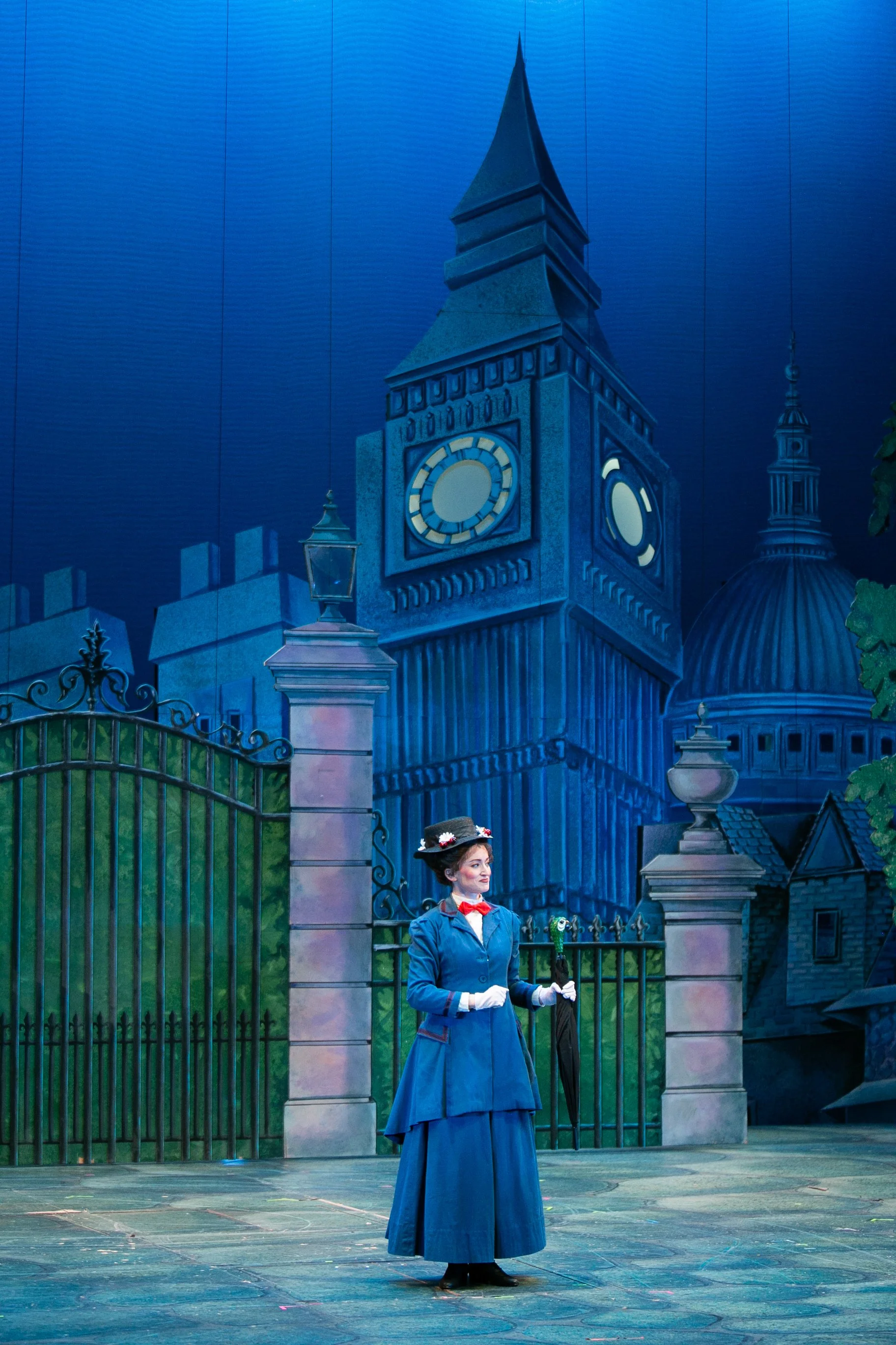 Woman dressed as Mary Poppins standing on stage in front of a painted background of Big Ben and a cityscape, holding a black umbrella with a green figure on top.