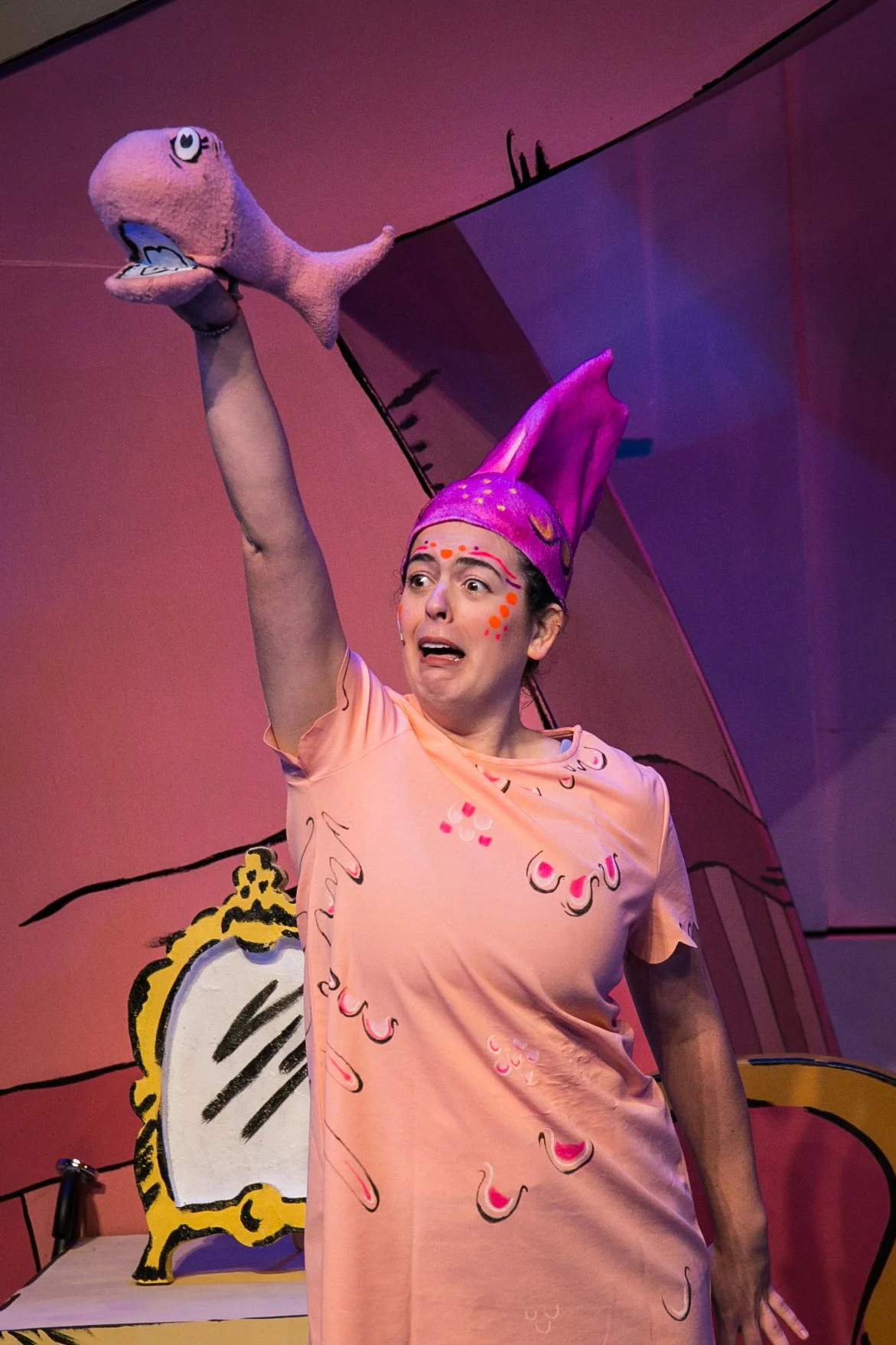 A woman dressed as a colorful character with a purple hat and pink dress, holding a pink fish puppet, performing on stage with a cartoonish set in the background.