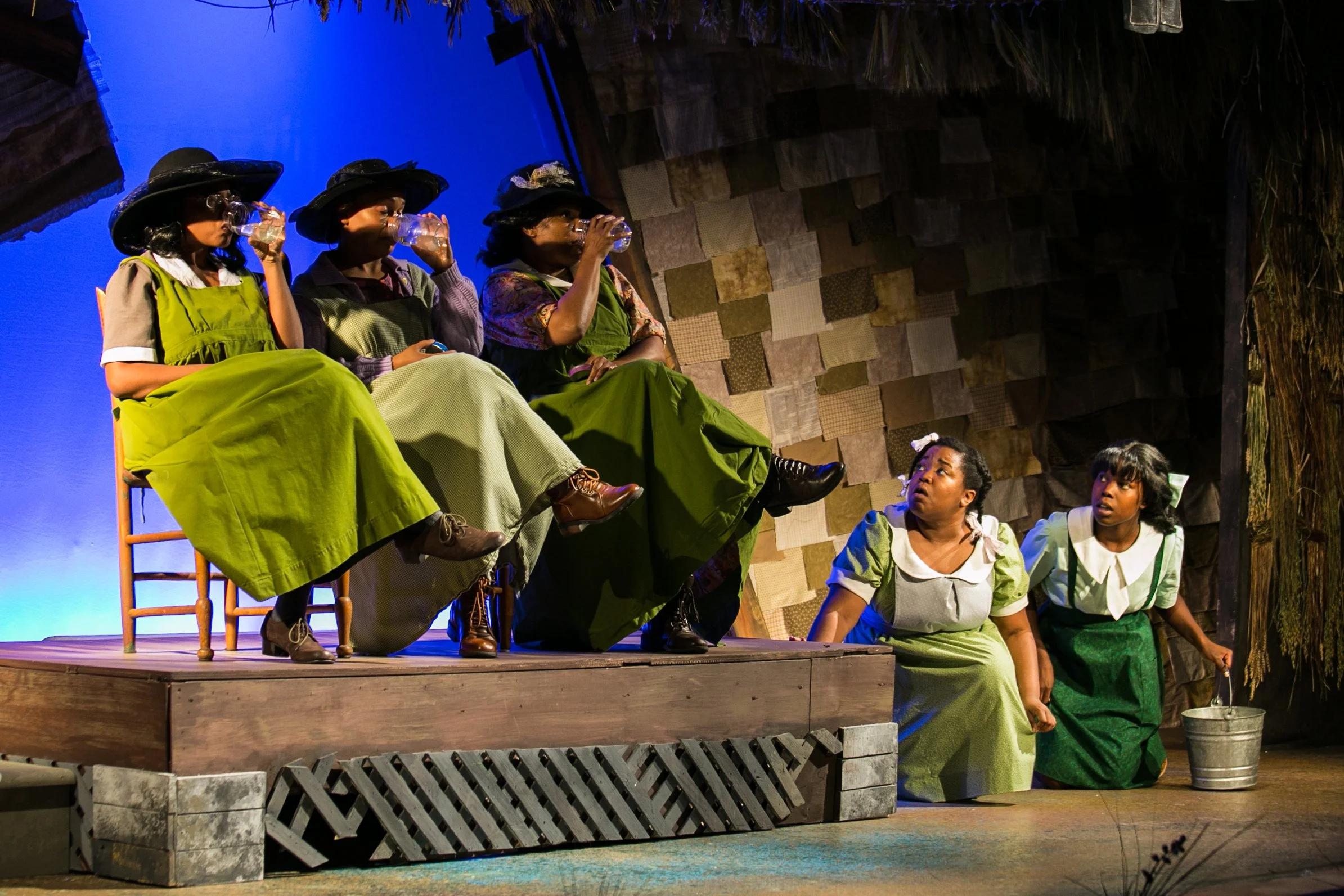 Group of women on stage during a theatrical performance, wearing vintage-style dresses and hats, with two women sitting on the floor. Some women are drinking from glasses, with a backdrop of textured wall and blue lighting.