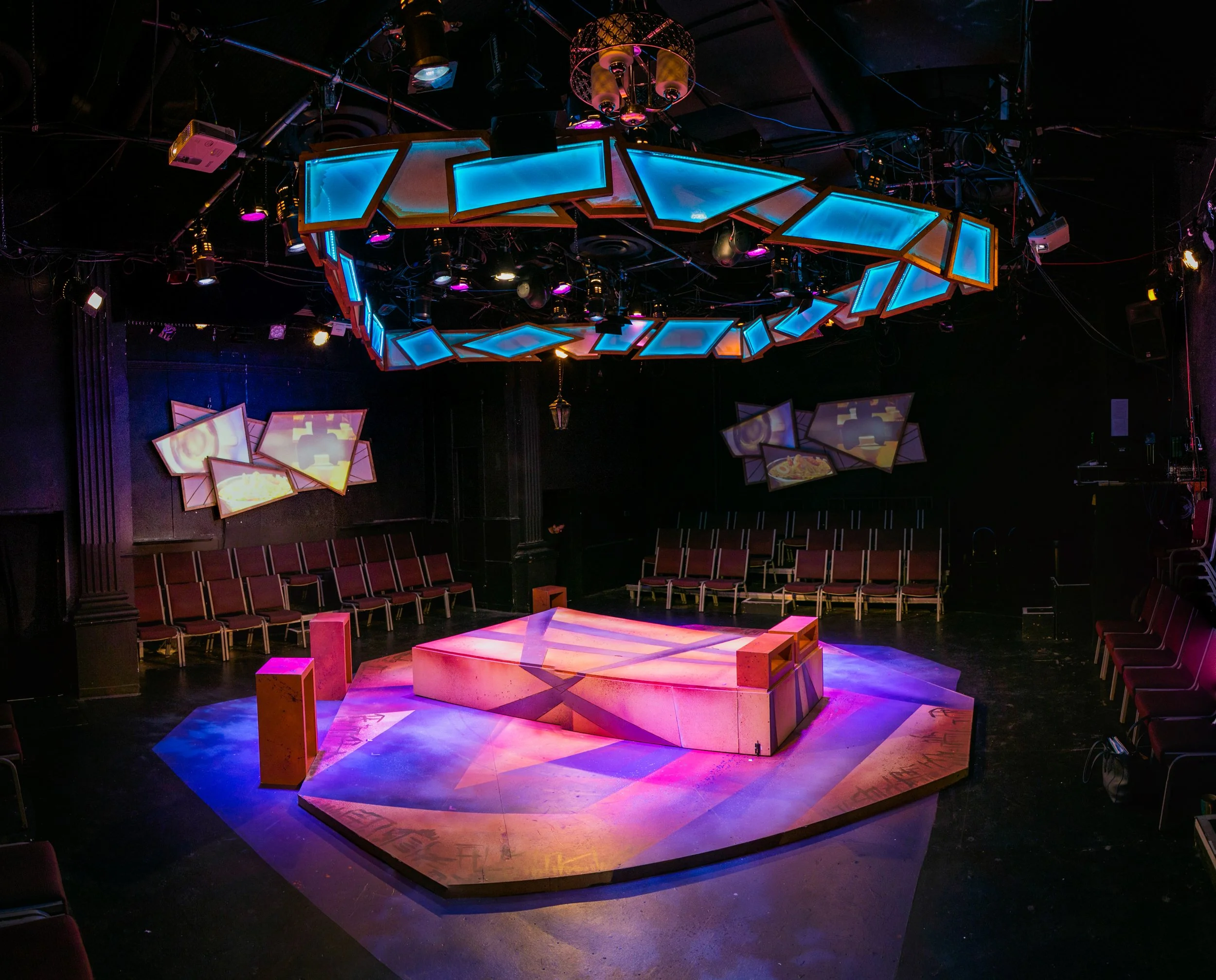 Empty stage with pink and purple lighting, surrounded by chairs arranged in a semi-circle, with abstract geometric light fixtures hanging from the ceiling in a dark room.