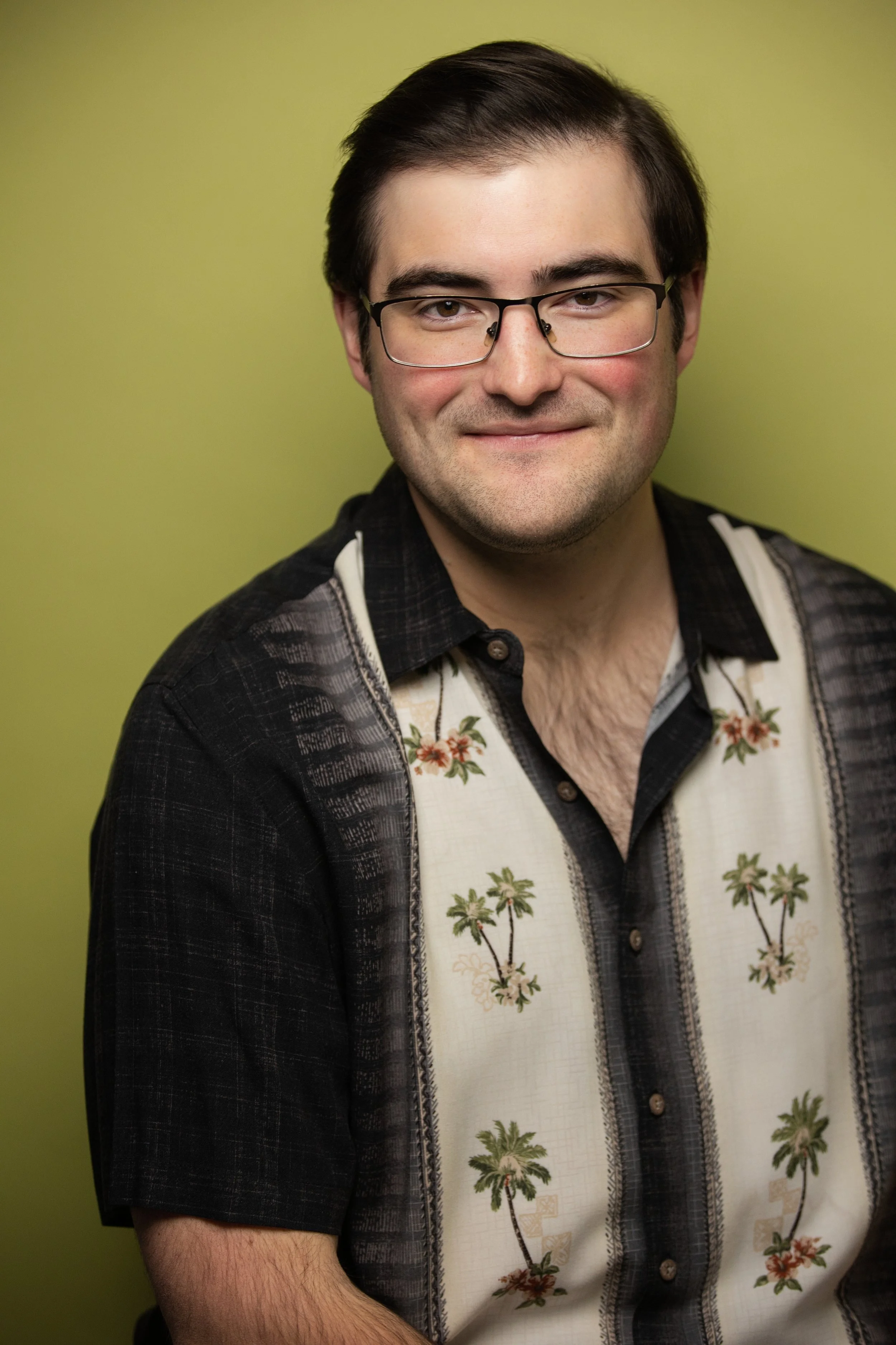 A young man with dark hair, glasses, and a slight smile, wearing a black and white Hawaiian shirt with palm trees, standing against a yellow-green background.