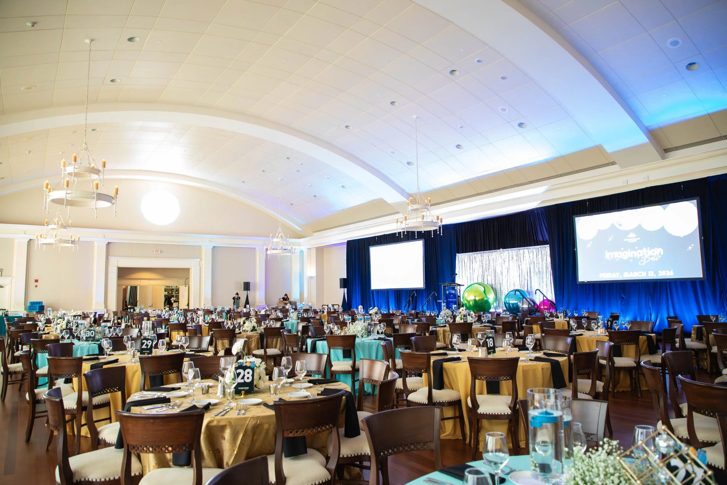 Spacious banquet hall decorated for an event with round tables, black and gold tablecloths, chairs with white cushions, large screens on stage, colorful decorations, chandeliers hanging from the ceiling, and blue lighting accents.