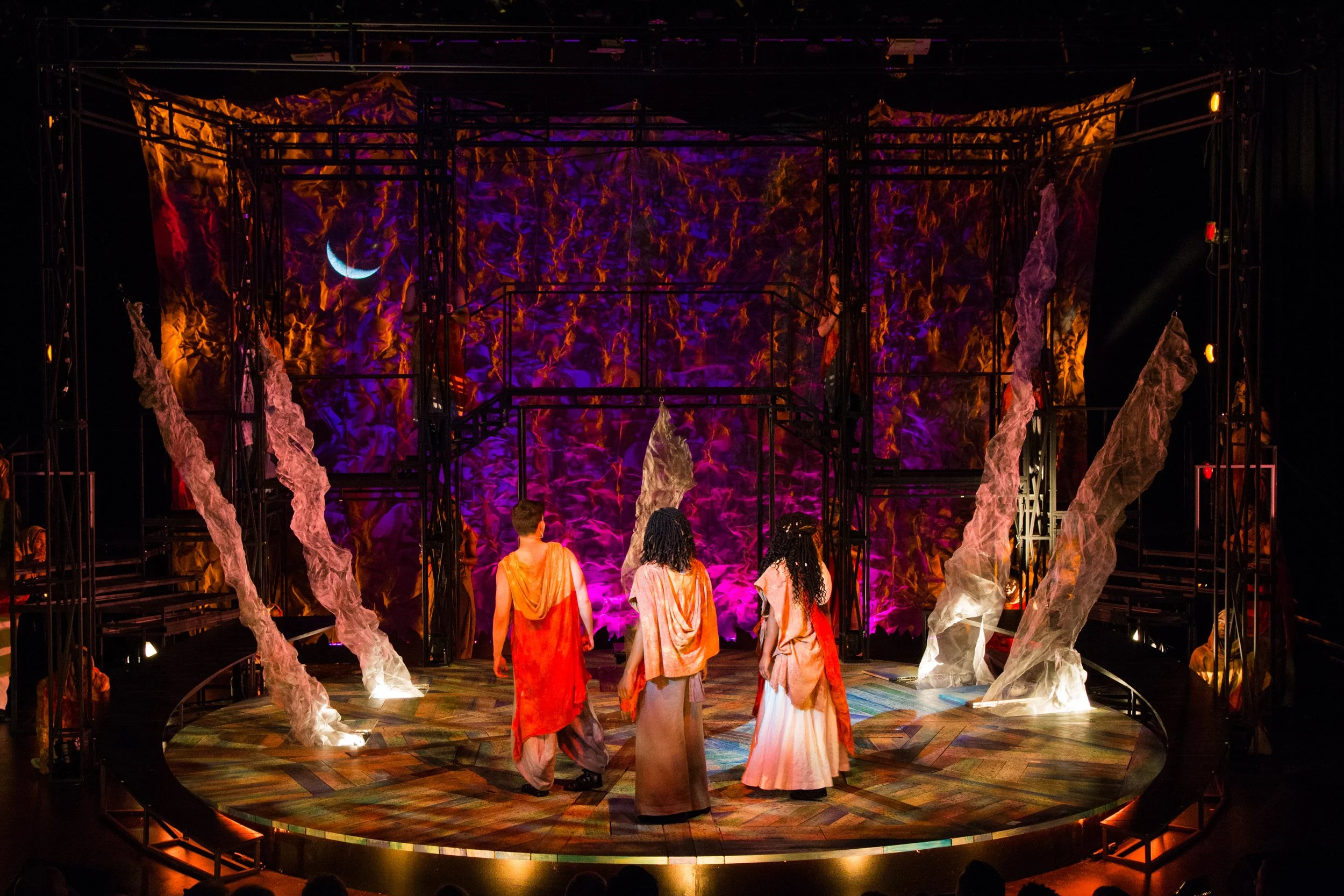 Theatre stage with four actors dressed in flowing costumes walking towards a backdrop showing a crescent moon and a starry sky, illuminated with purple and orange lighting.