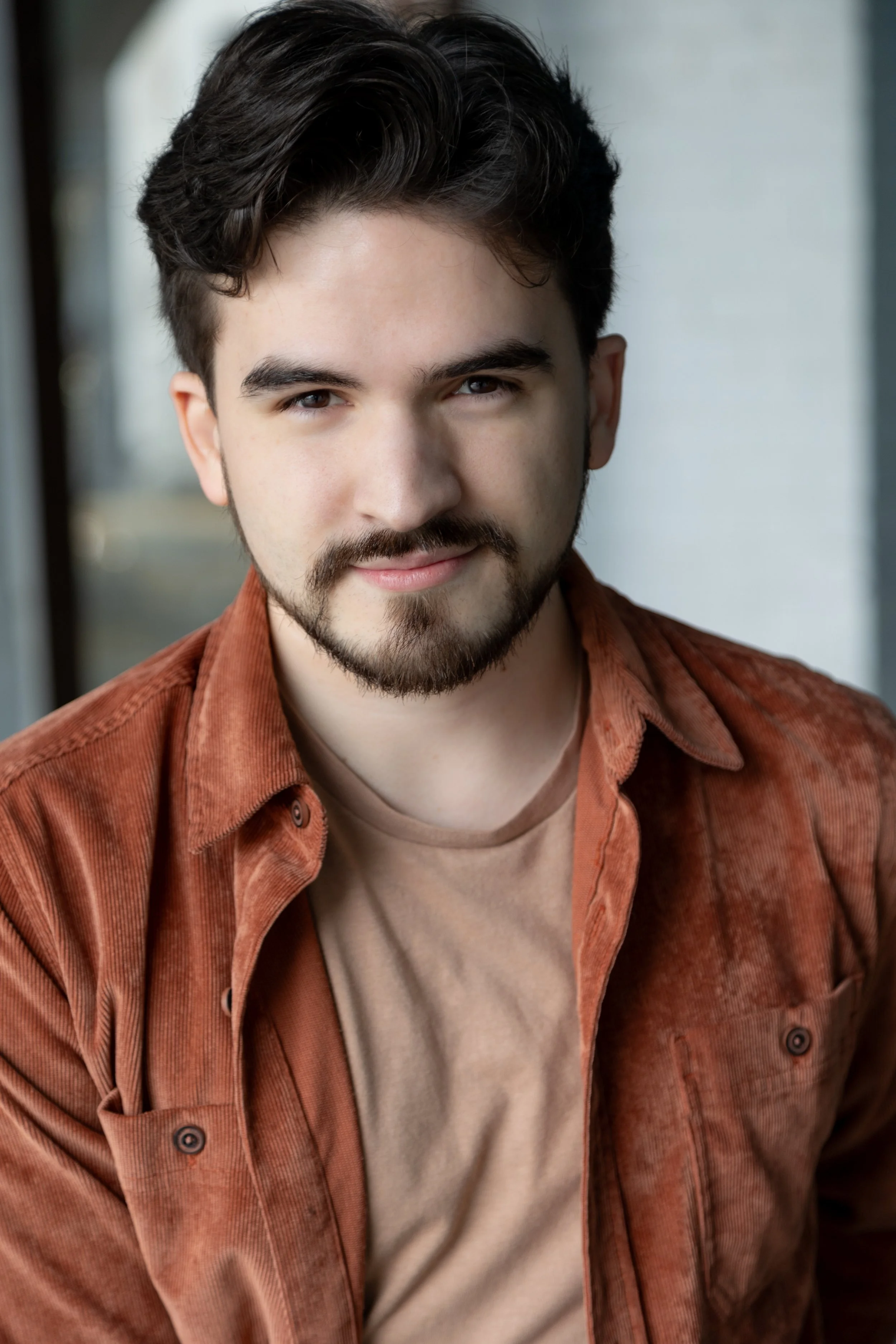 A young man with dark hair and a beard smiling, wearing a brown jacket over a beige shirt, in an indoor setting with a blurred background.