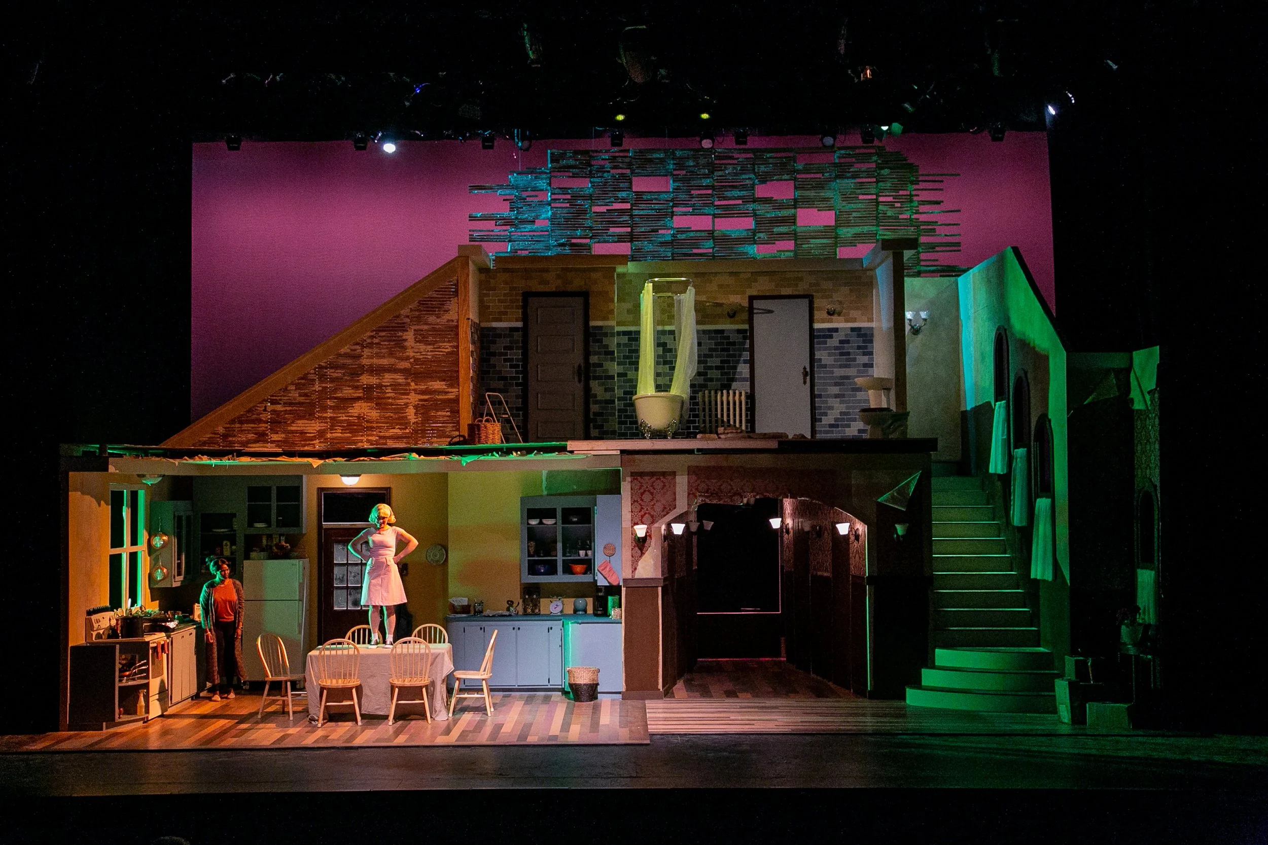 A theatrical stage set depicting a two-story house interior with furniture, characters, and colorful lighting.