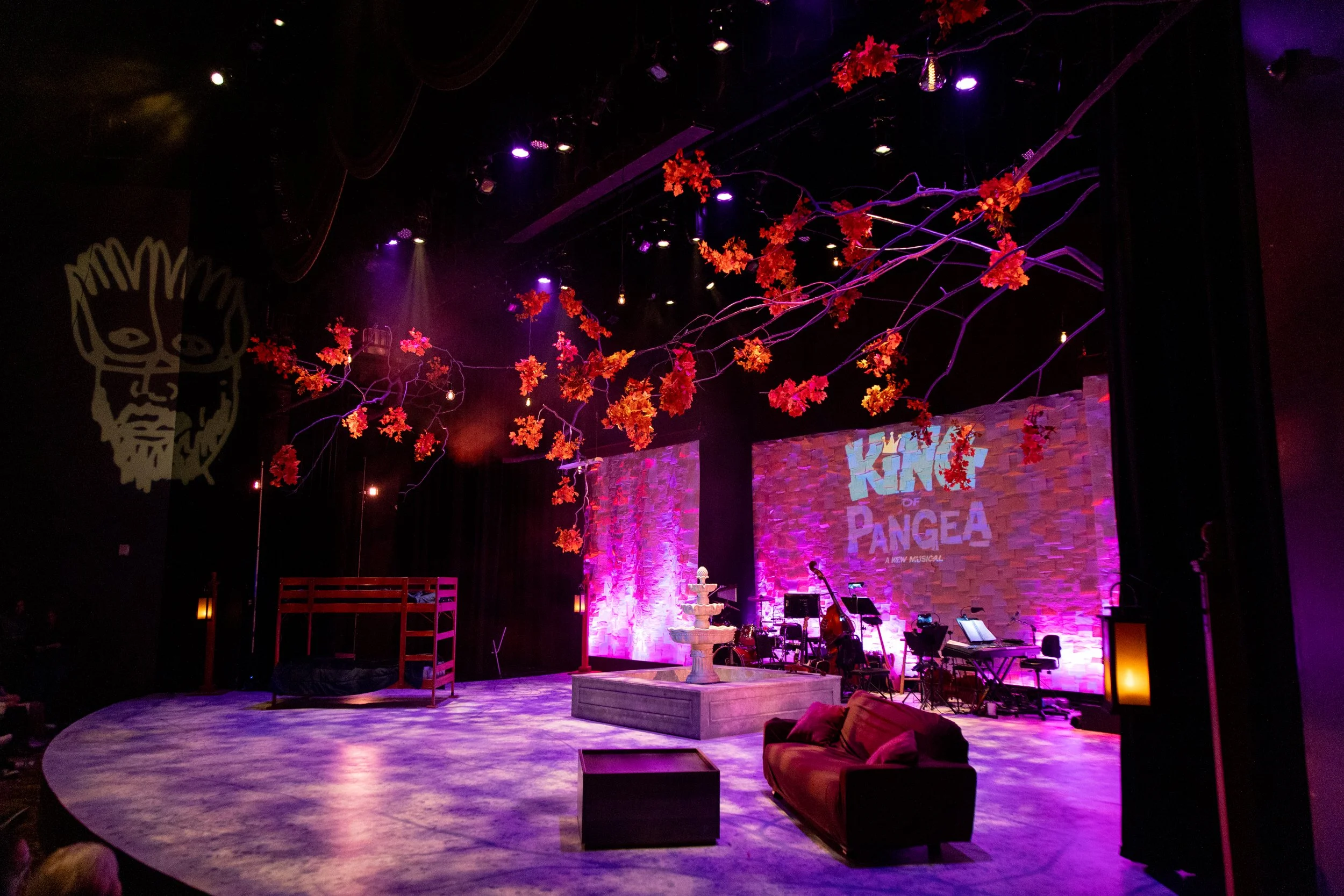 The stage is set with a backdrop reading "King of Pangea, A New Musical" illuminated in purple lighting. Overhead, pink and orange leaves and branches hang, creating a decorative canopy. The stage features a fountain, a red bunk bed, a brown sofa, an