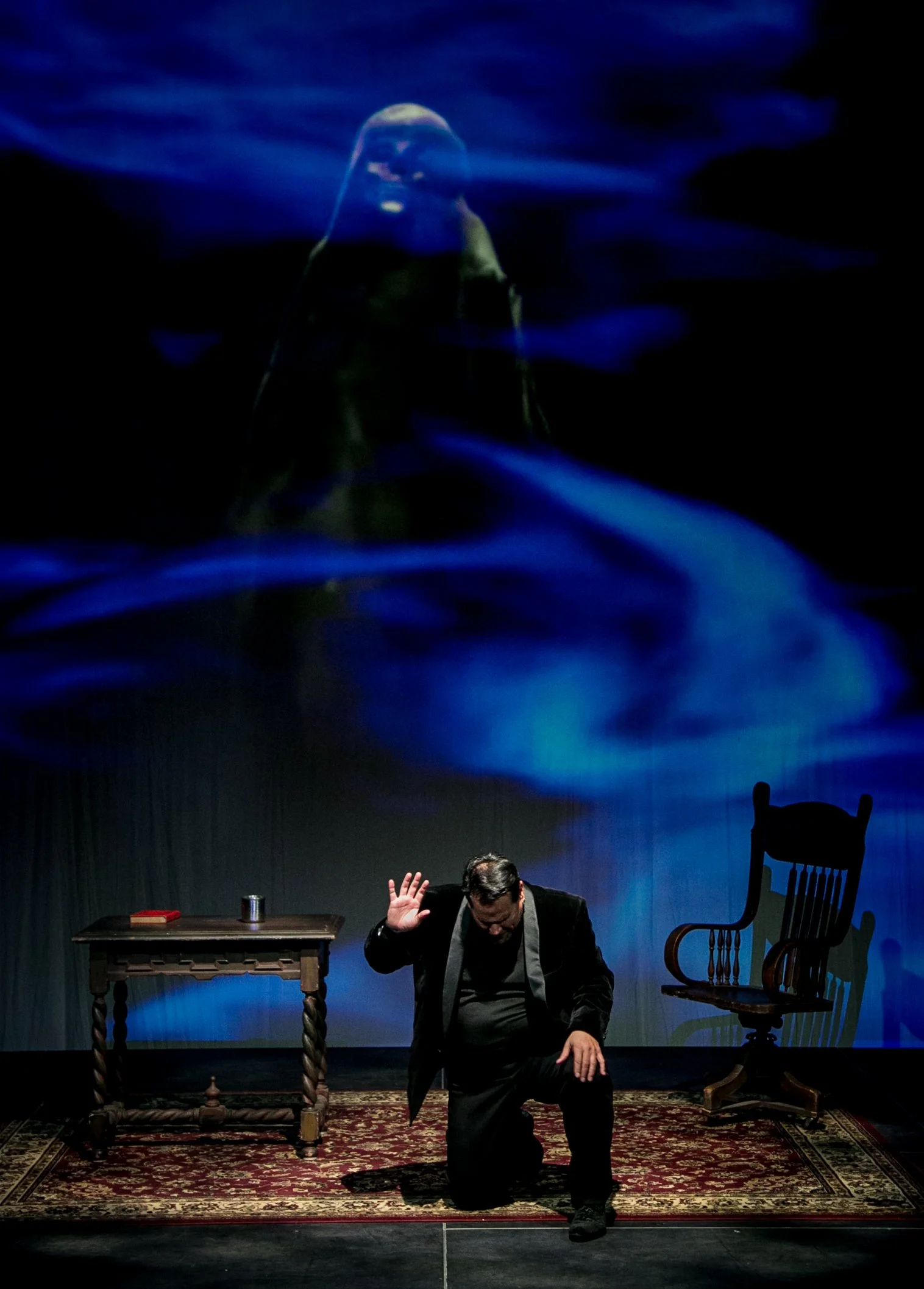 A man kneeling on stage with a hand raised, performing in front of a dark background with a large projection of a woman’s face and blue abstract patterns. A wooden table with a book and an ashtray, and a wooden rocking chair are also on stage.