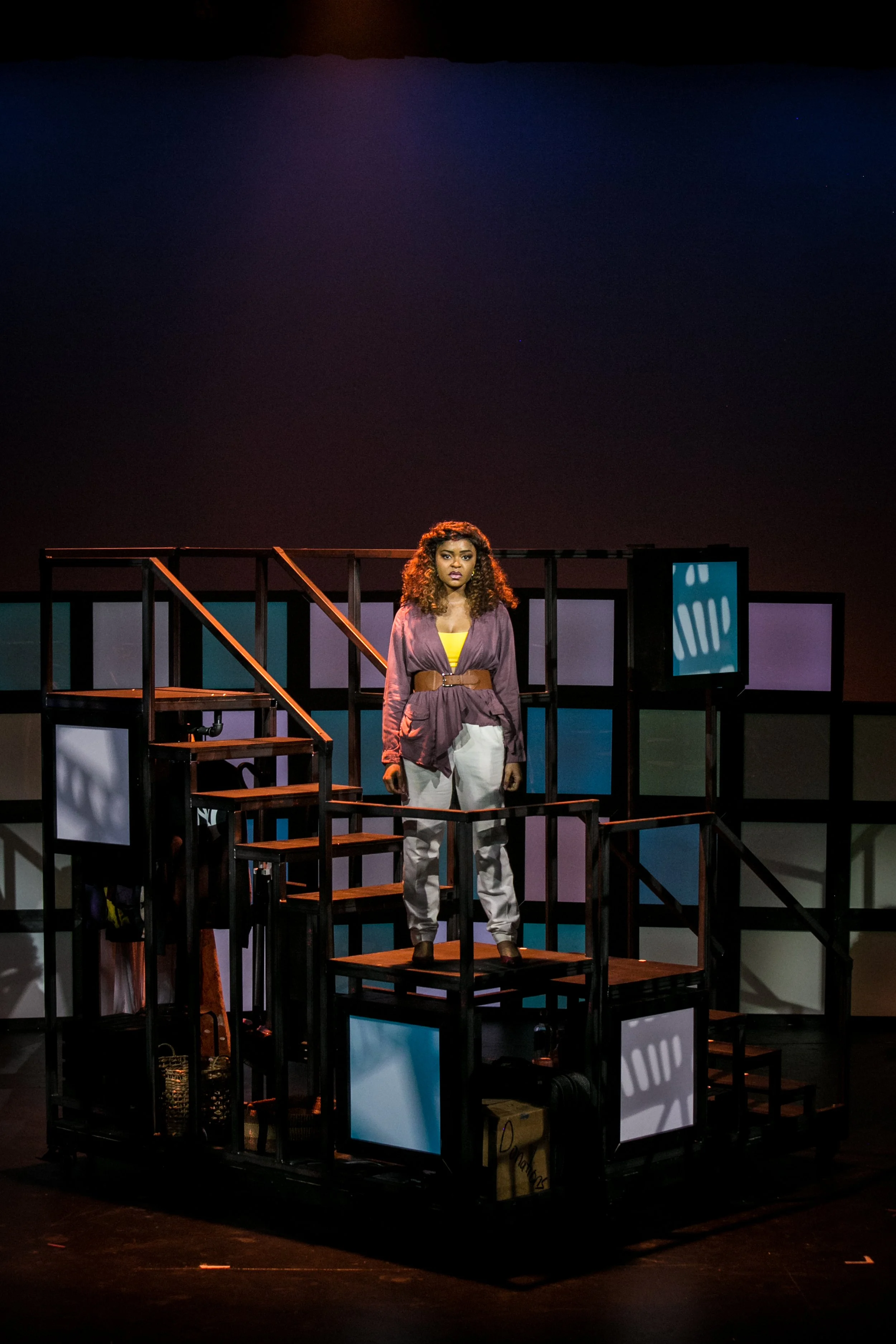A woman standing on a multi-level stage set with colorful framed panels in the background, likely performing in a theatrical production.