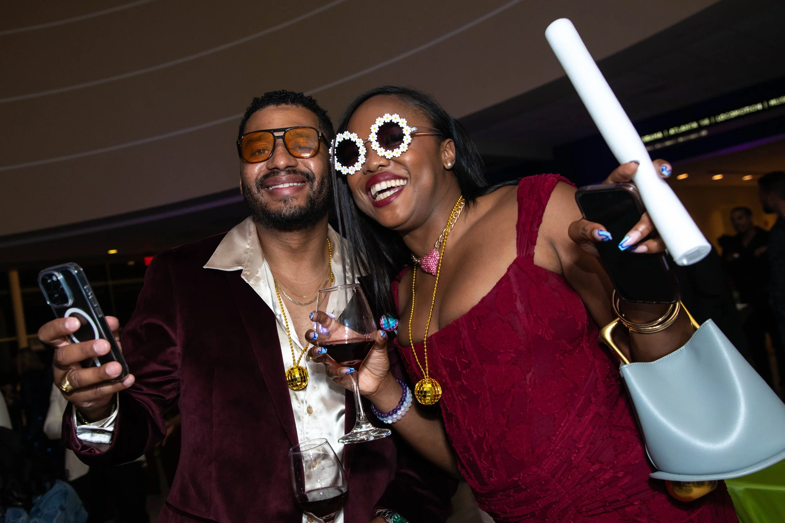 A man and woman dressed in party attire at a celebration, both holding drinks and taking selfies, wearing colorful sunglasses and festive accessories.
