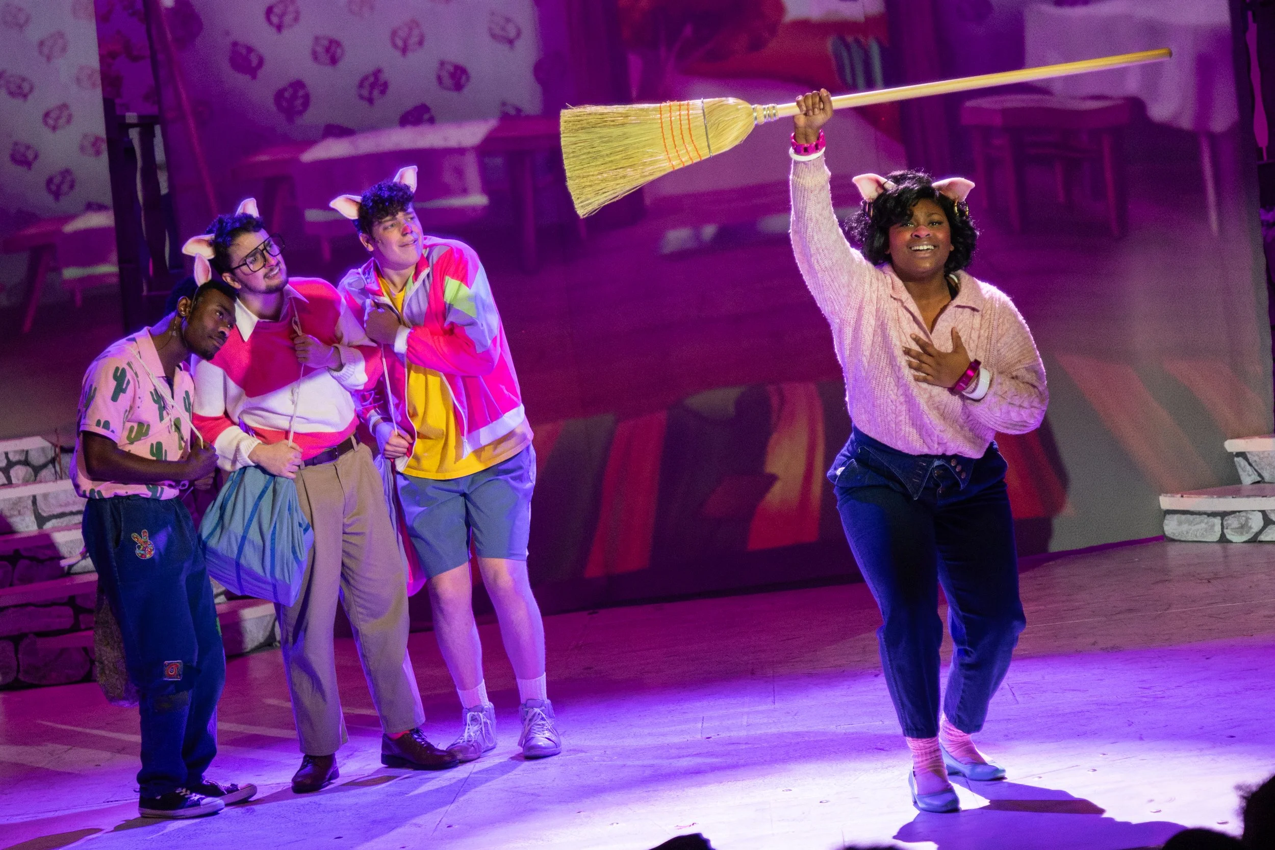 A theatrical stage scene with four performers dressed in colorful costumes with animal ears and accessories. One woman is holding a broom and appears to be singing or speaking passionately, with the other three looking at her, smiling and appearing a