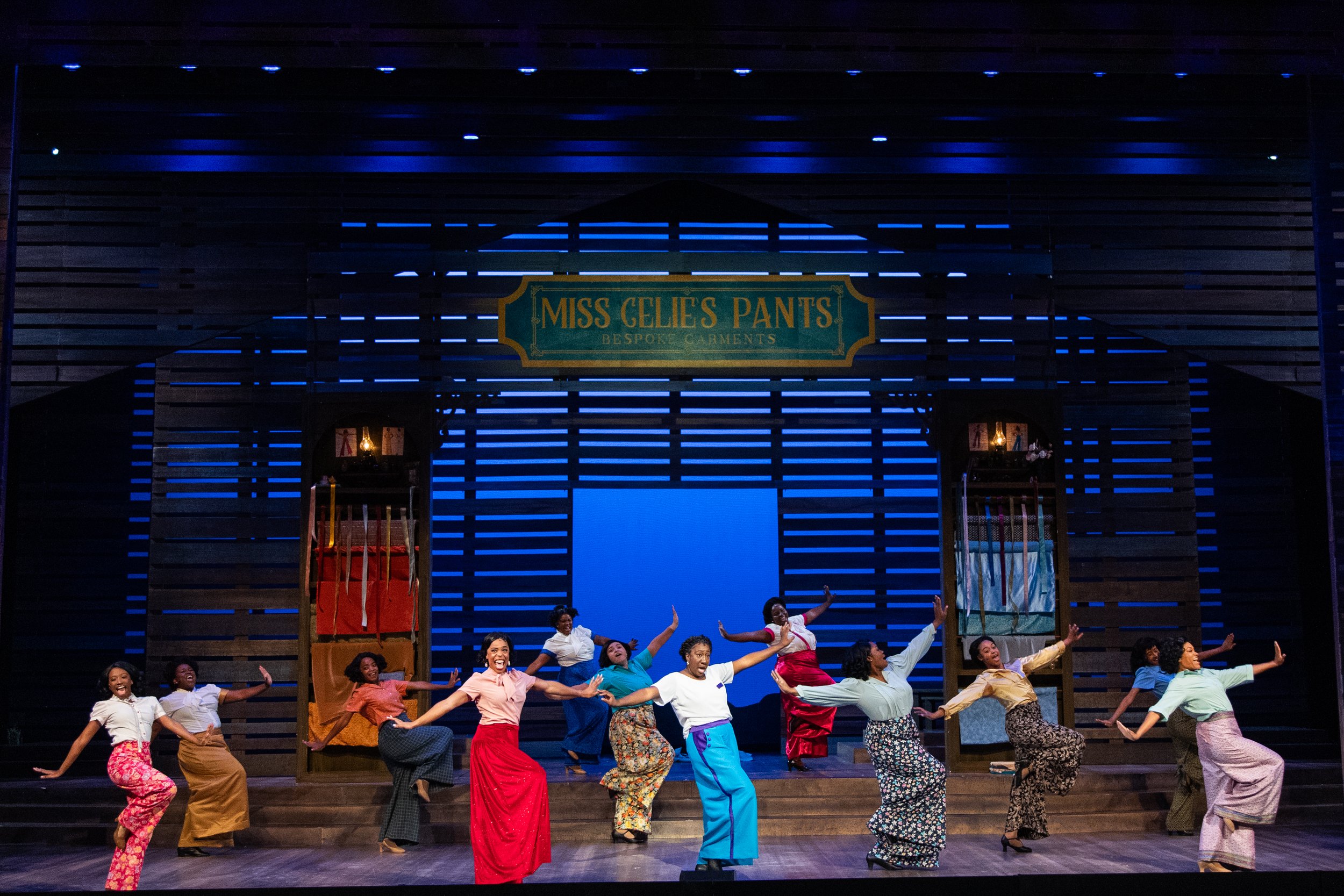 Stage performance with dancers in colorful skirts and blouses, in front of a rustic set with a sign that reads "Miss Celie's Pants Bespoke Garments"; group dancing with arms raised.