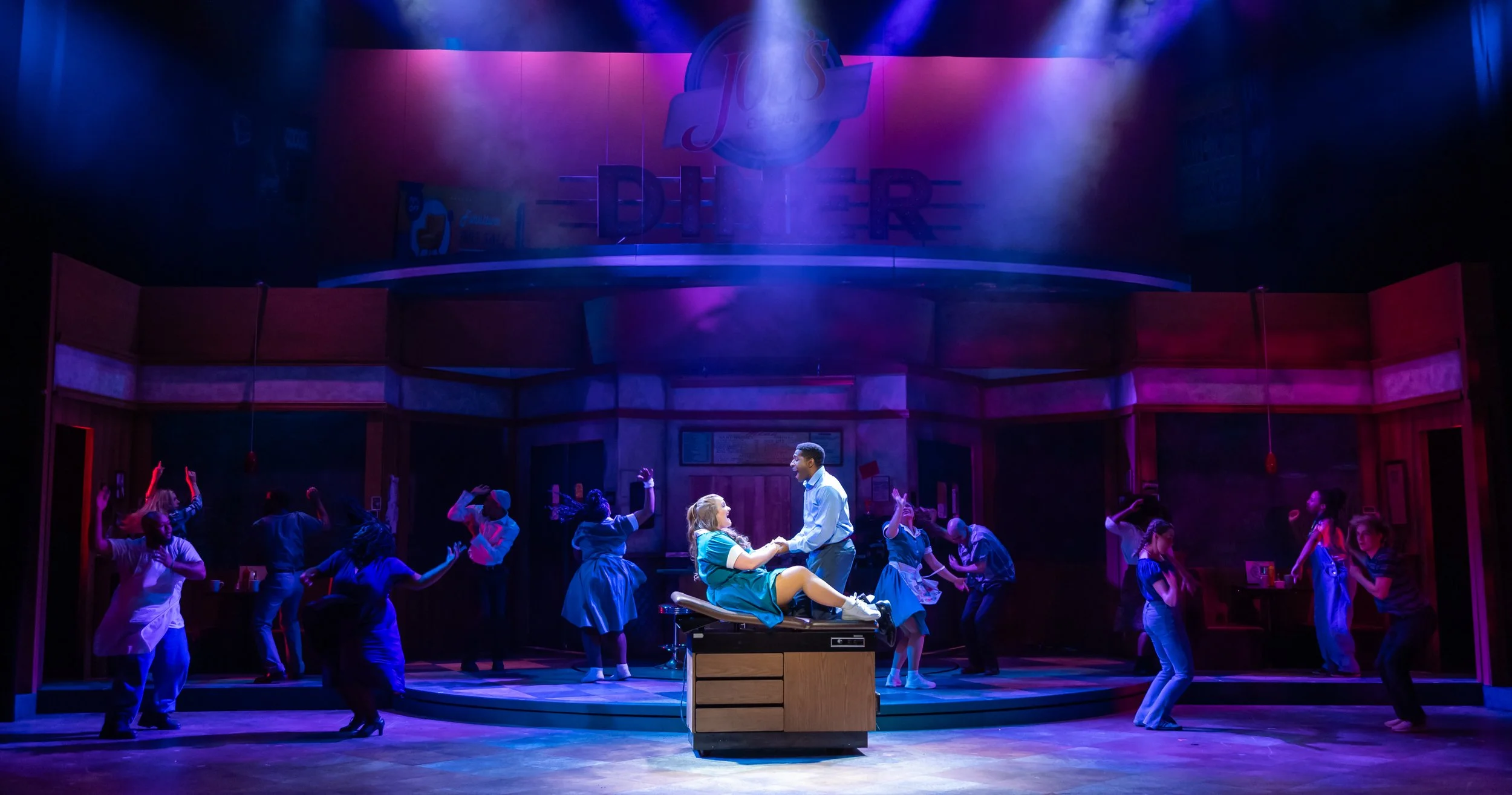 Stage scene from musical 'Diner,' with performers dancing and singing in colorful lighting, center features two actors in a close embrace.