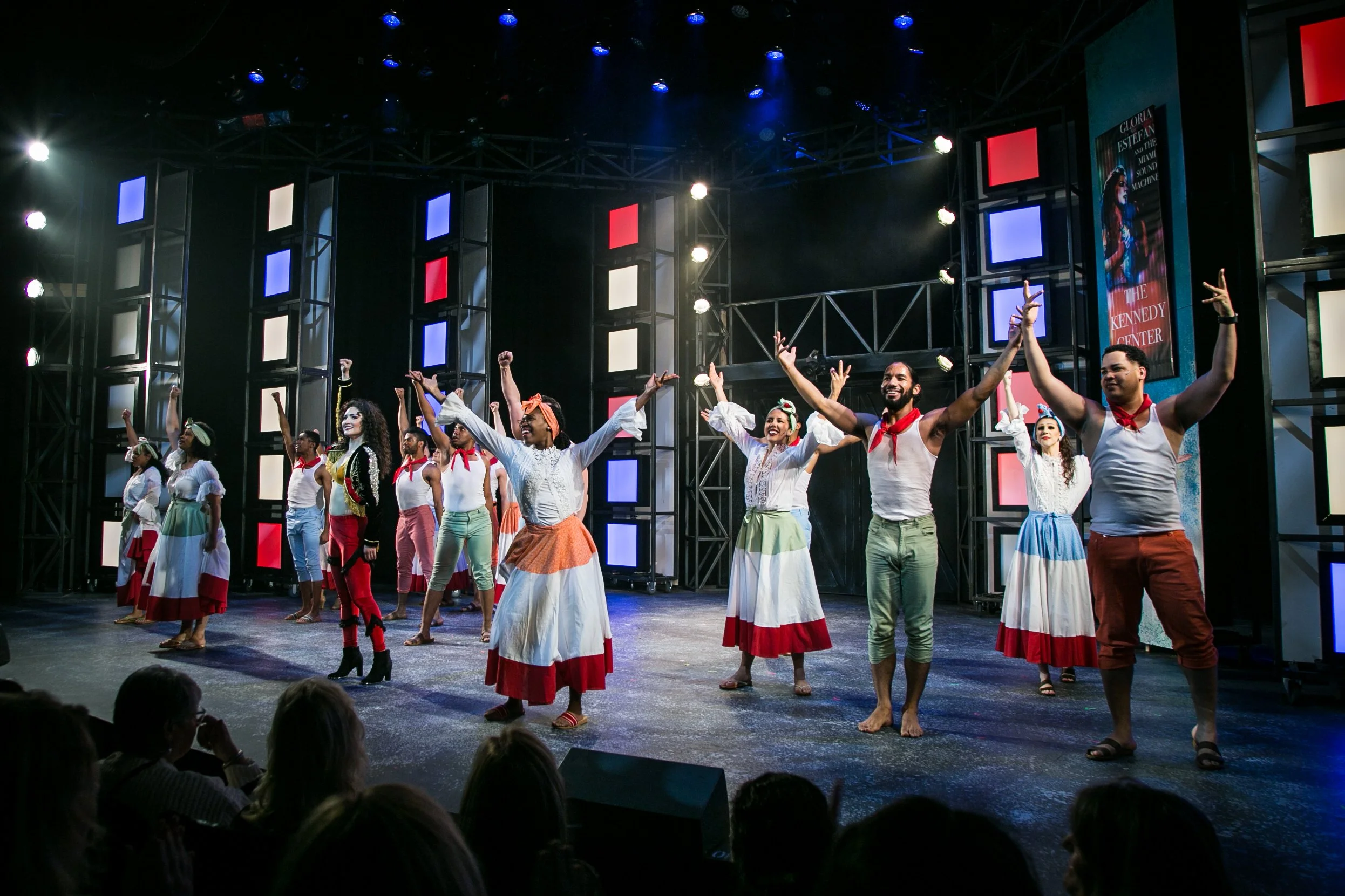 The image shows a group of performers on stage during a theatrical performance. They are dressed in colorful costumes, some with skirts and headscarves, and are raising their hands in a celebratory manner. The stage has vibrant lighting with blue, re