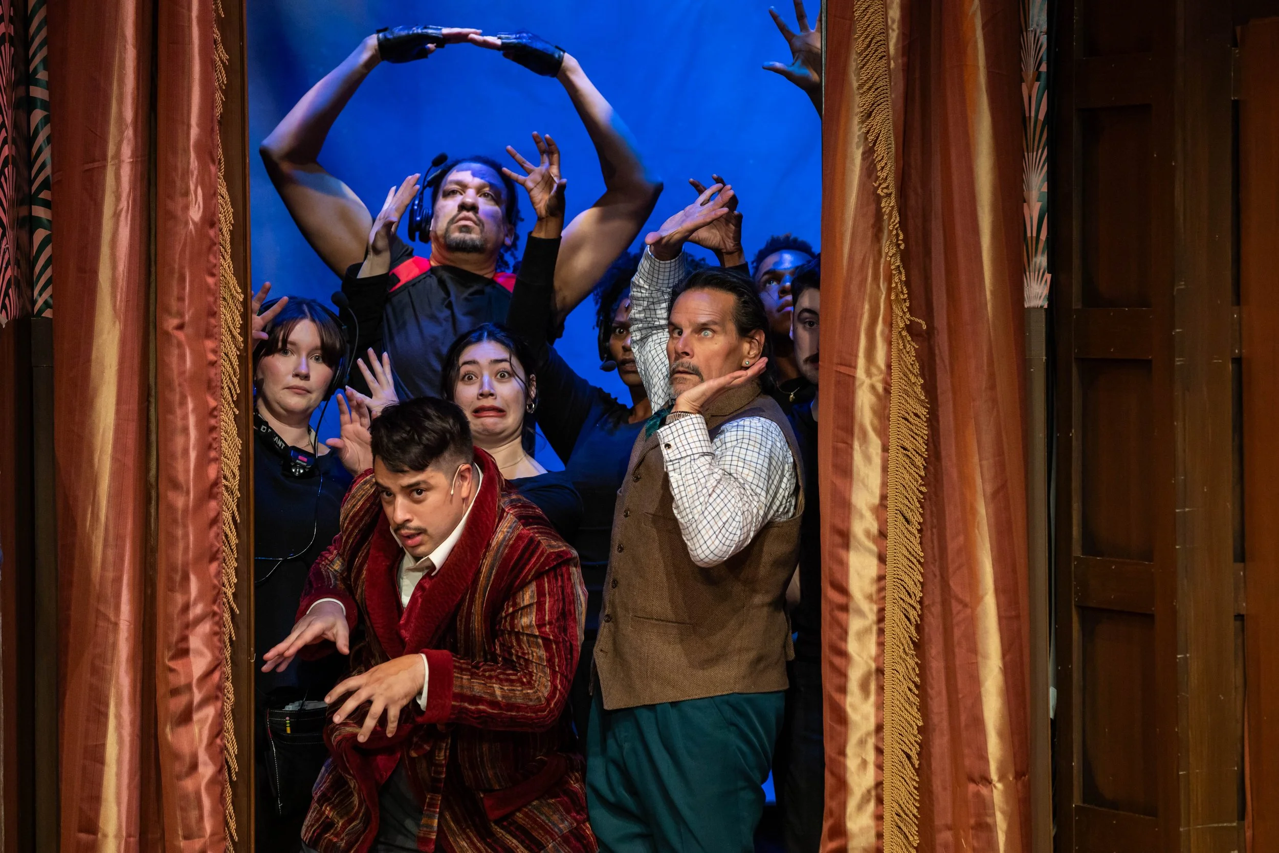 A group of people on stage appearing surprised or frightened, framed by red velvet curtains, with a blue background.